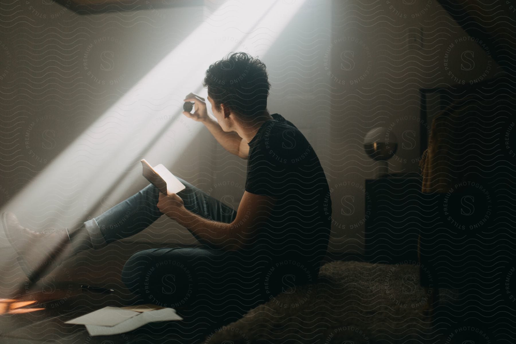 Young man reading a book while sitting on the floor of a room holding a coin under morning light