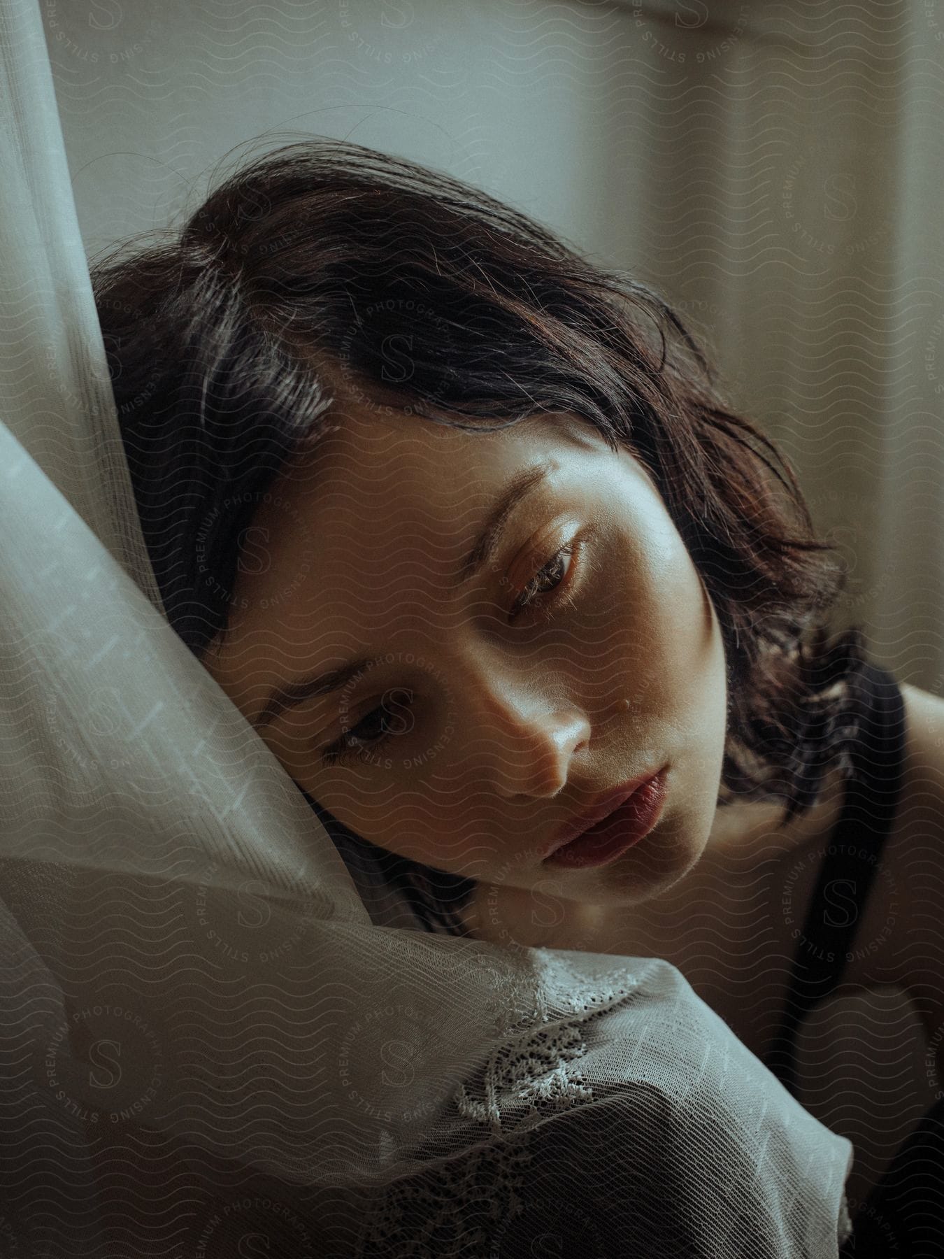 A brunette woman with a somber expression wearing a black strap top leans against a curtain