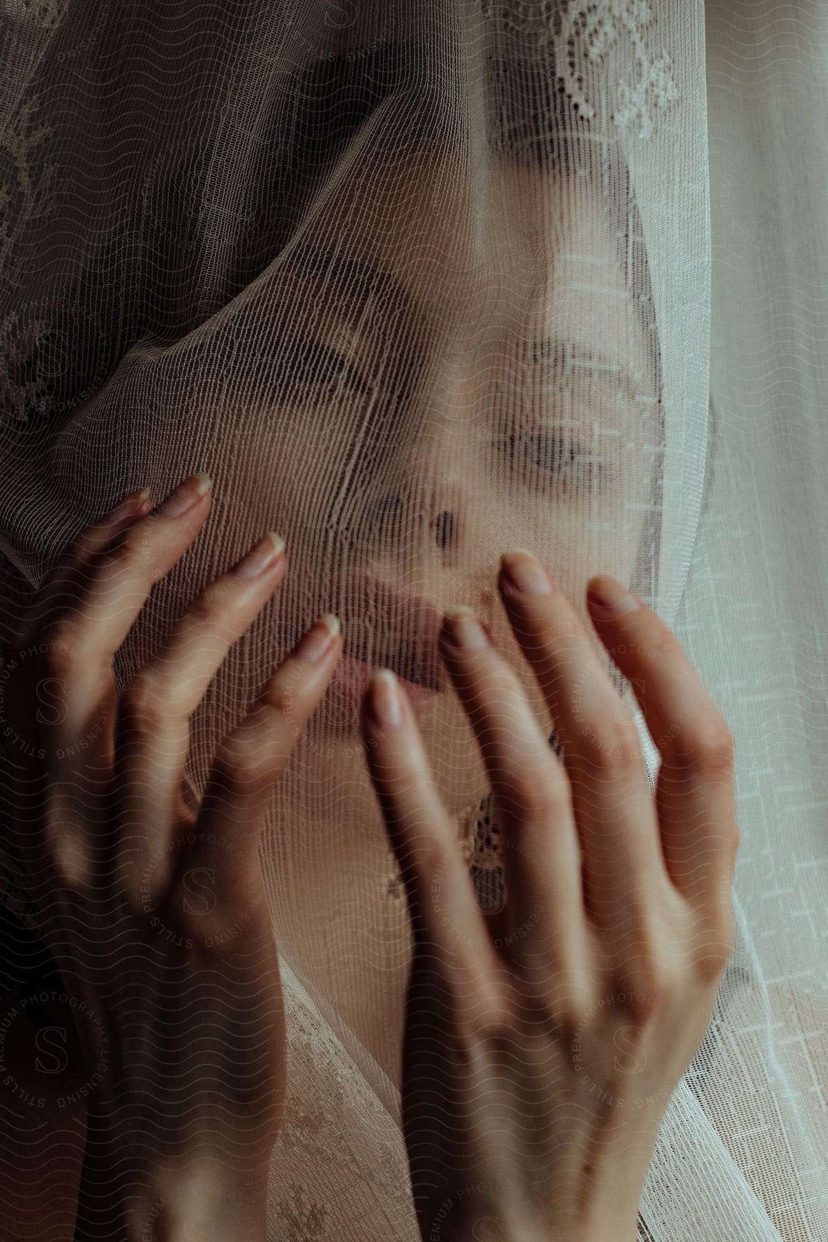 A woman posing with her hands in front of her face covered by a lace blind