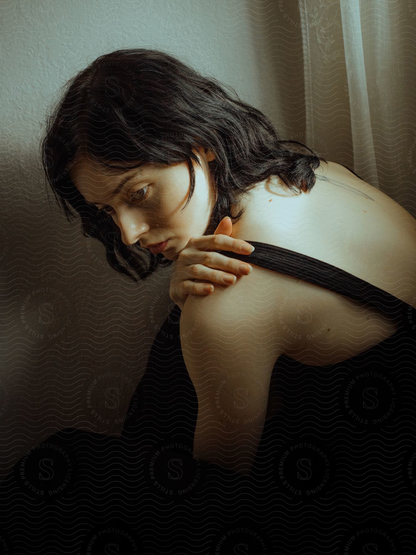 A darkhaired woman wearing black sits down and touches her shoulder next to a window in an interior setting