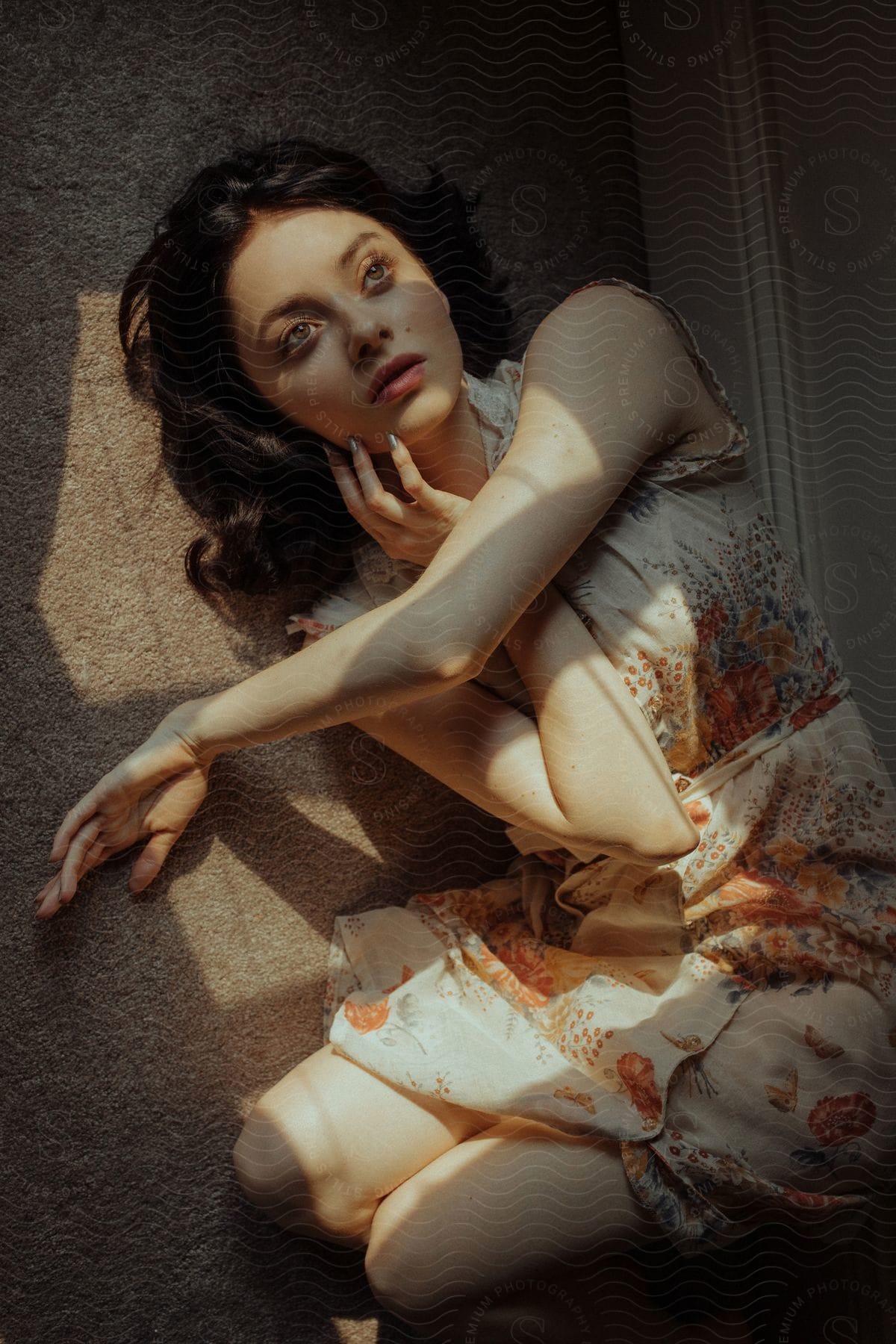 Stock photo of young woman in vintage flowered dress poses with arm across chest and another on face lying on tan carpet with shadow
