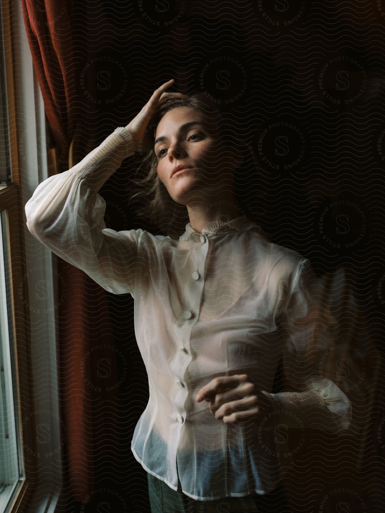 Young elegant woman wearing a translucent blouse looking out the window of a room