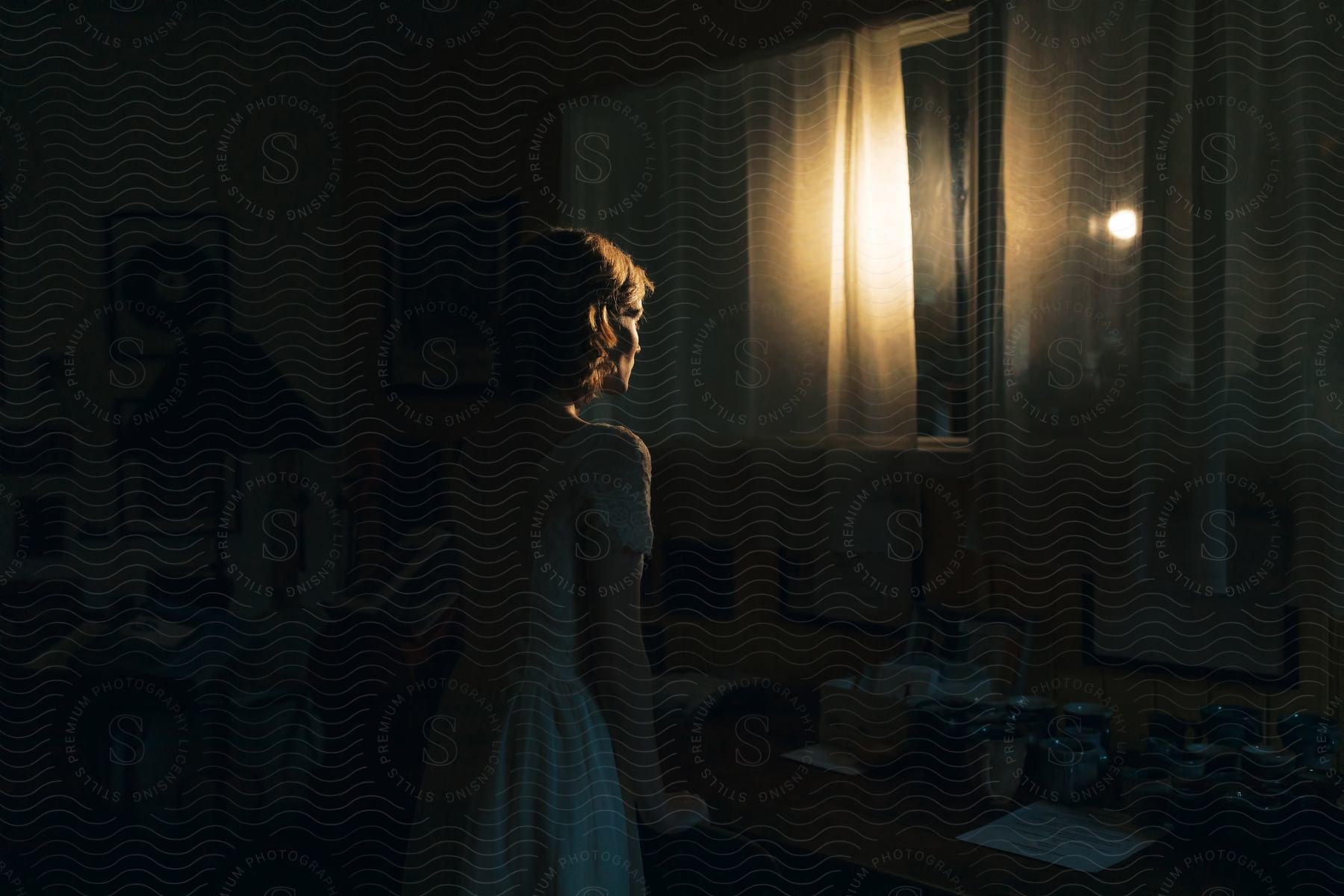 A woman wearing a white dress stands next to a table near a window seen from the back