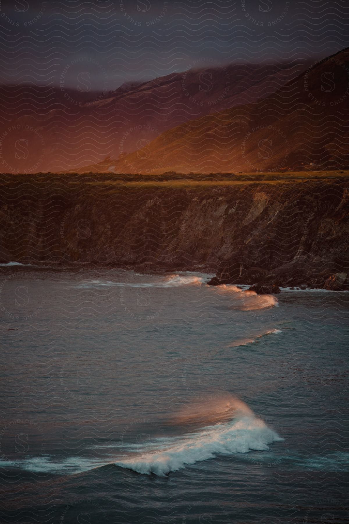 Ocean waves roll in at sunset with cliffs and green mountains in the background