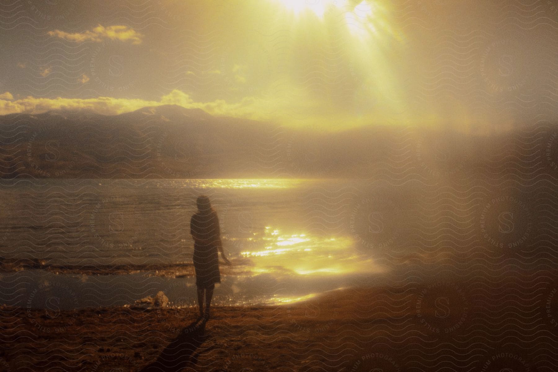 A woman stands on the shore of a small lake before mountains at dawn