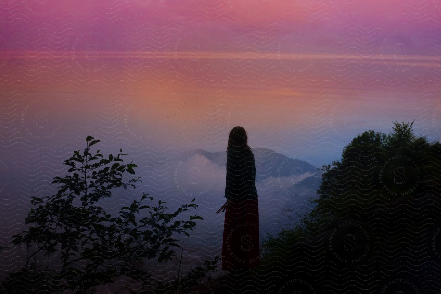 A Woman Standing On A Hillside Between Two Bushes Facing The Mountain And Colorful Sky With Her Hand On Her Side