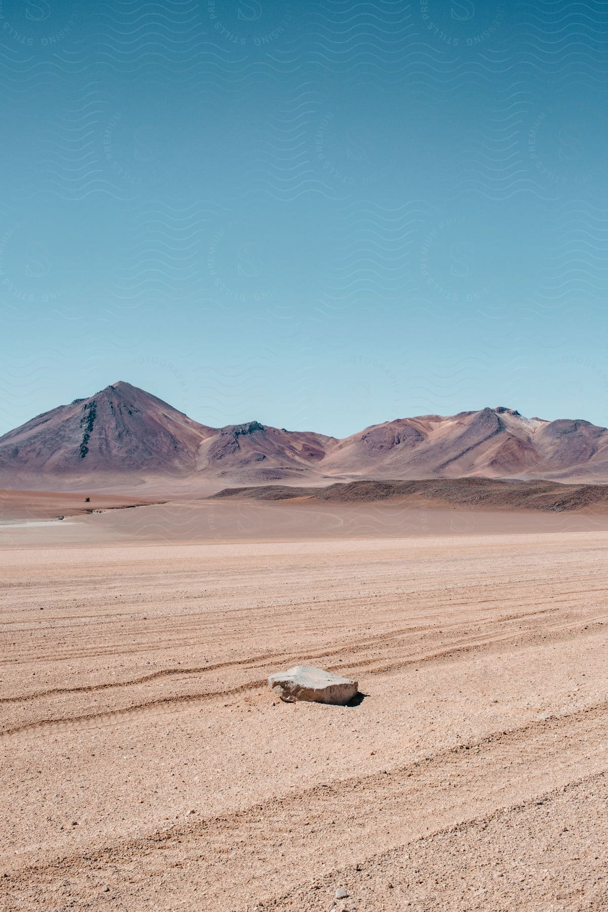 Solitary rock on desert sand next to mountains under a blue sky