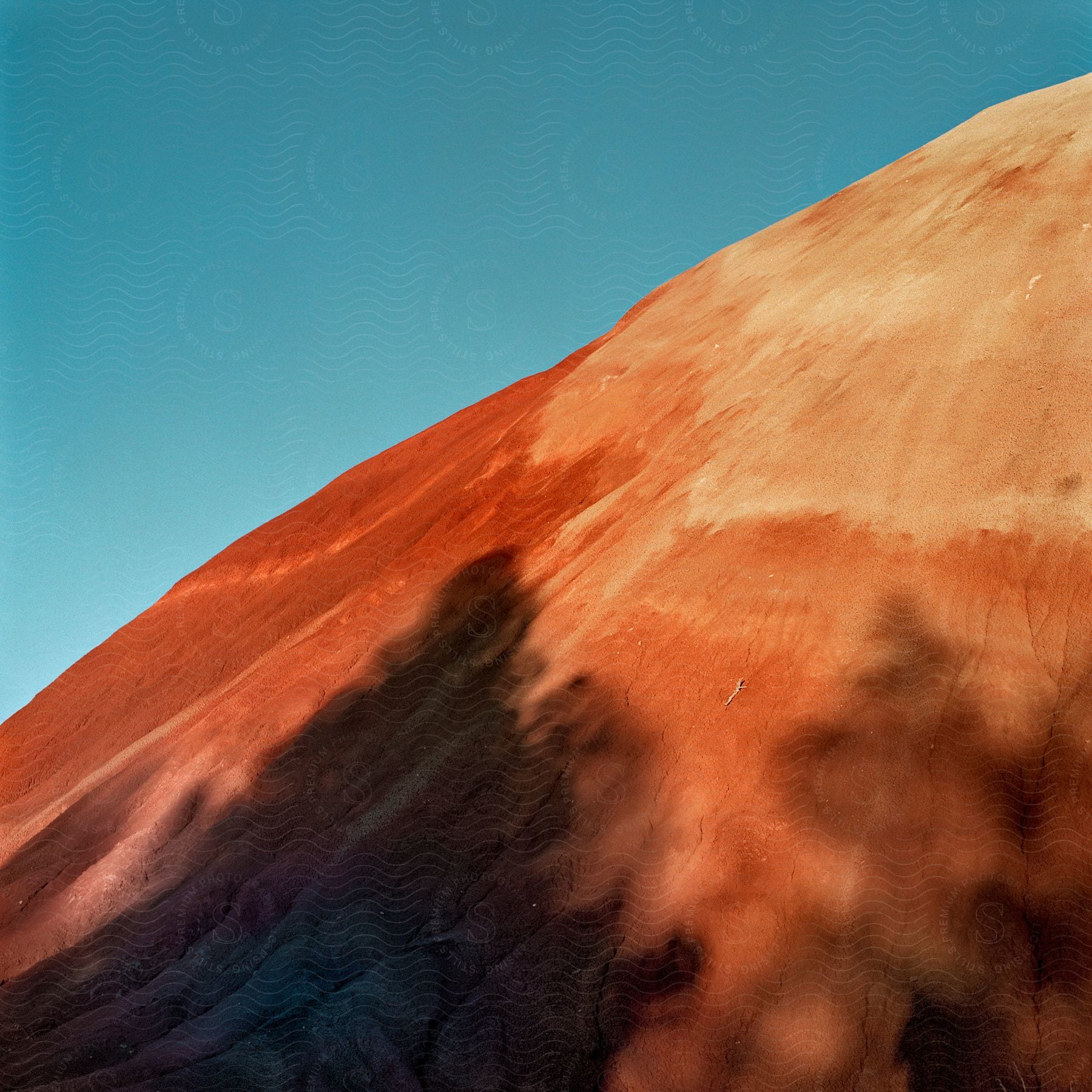 Tree shadows cast over orange slopes of a desert mountain under a blue sky