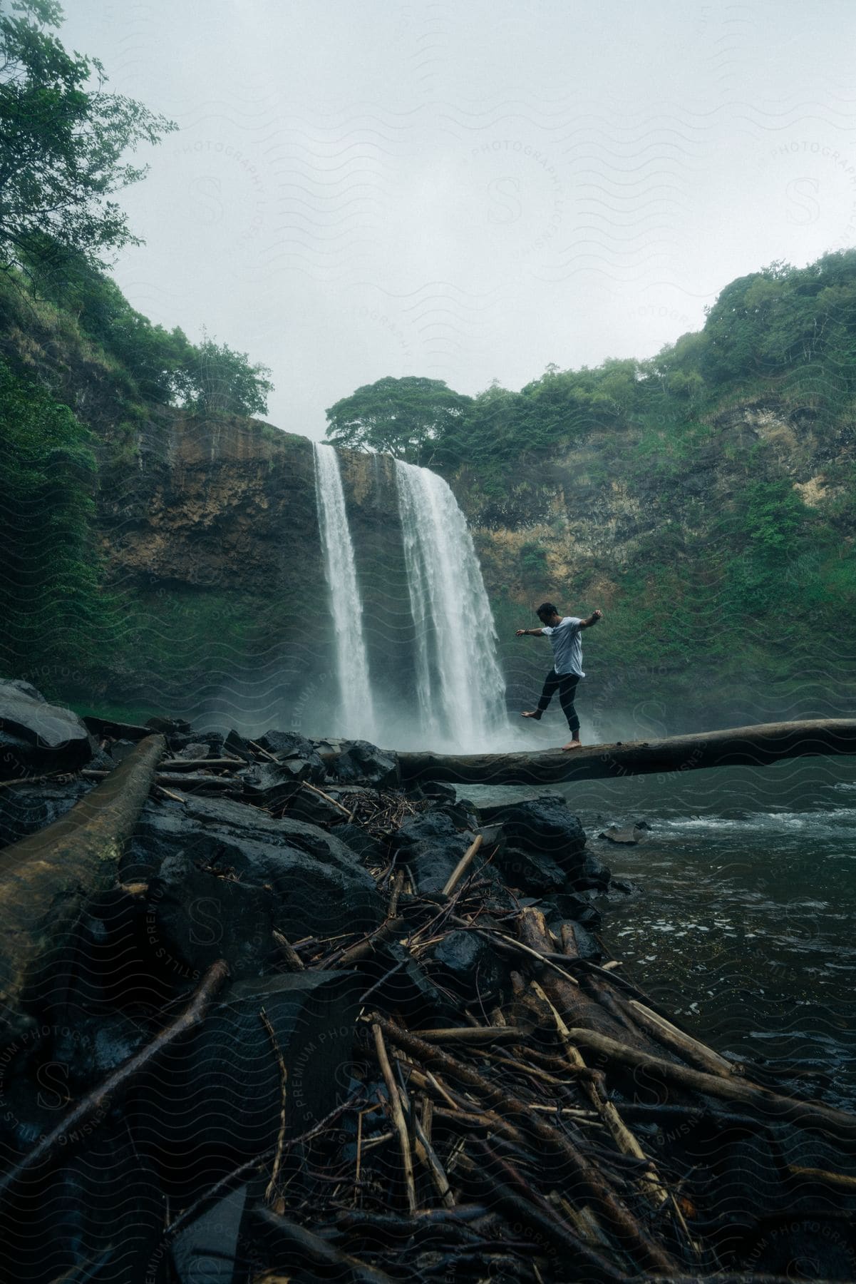 A young adult man balances on a fallen tree trunk over a river in front of a waterfall