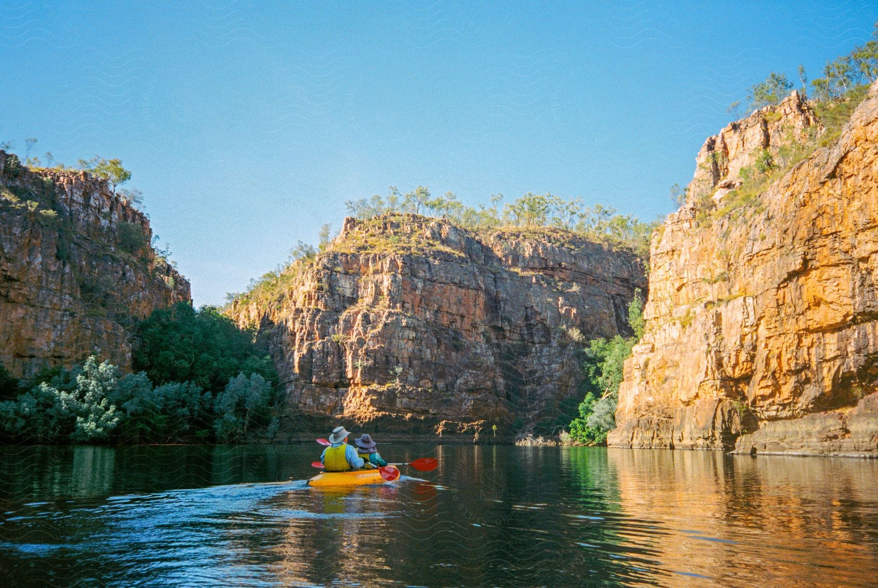 Two people kayaking down a river surrounded by high mountain cliffs with trees in the crevices