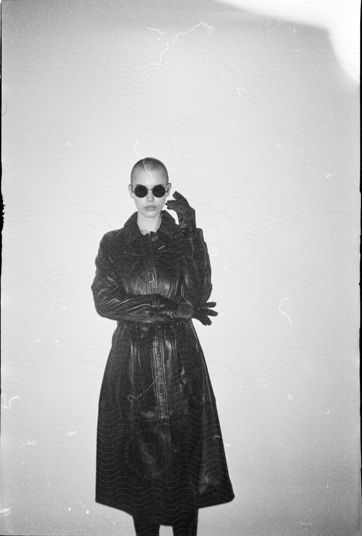 Woman with shaved head sunglasses and a black leather jacket stands in front of wall