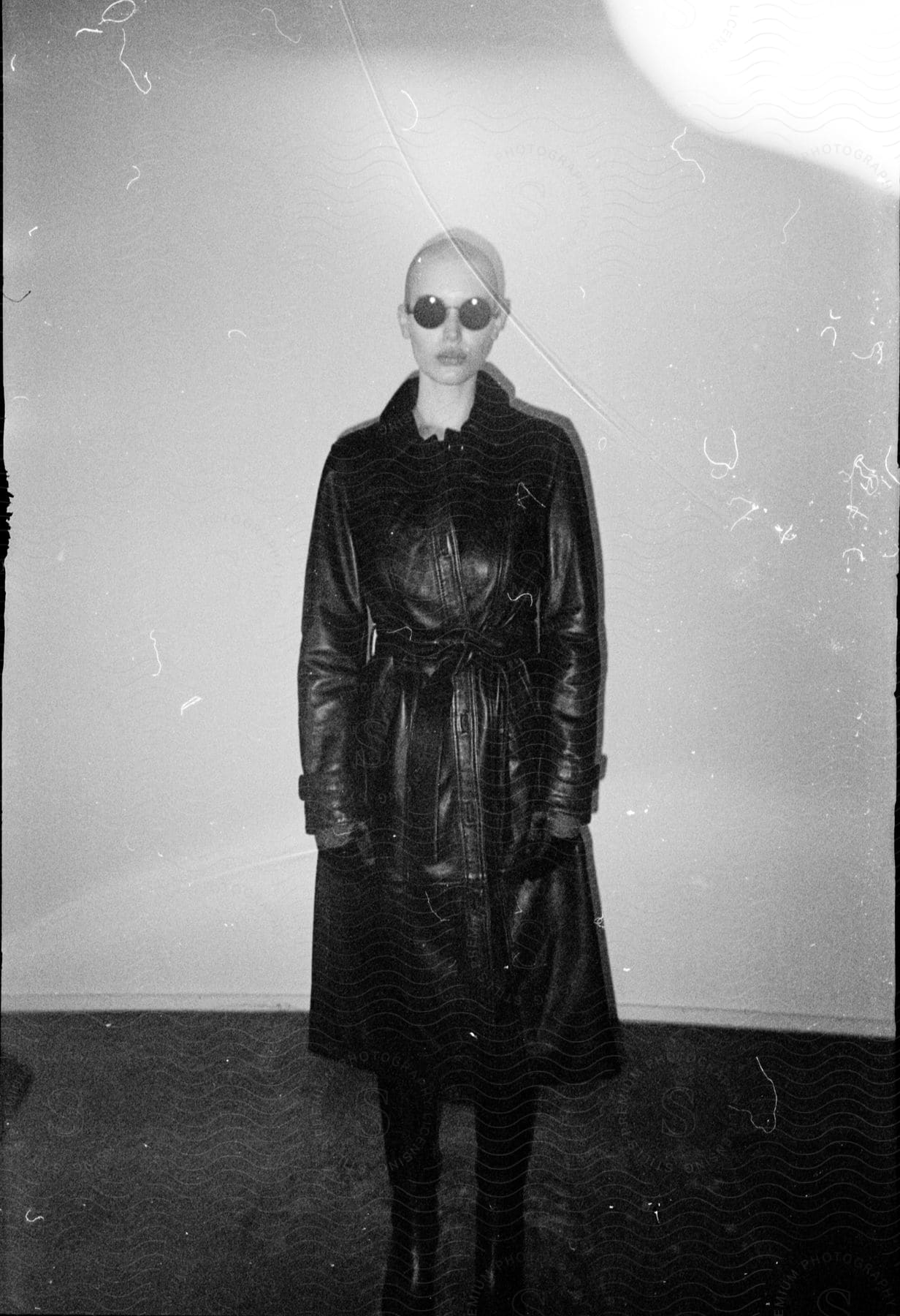 Woman with a shaved head, glasses, and a long black leather overcoat stands confidently in a dimly lit room