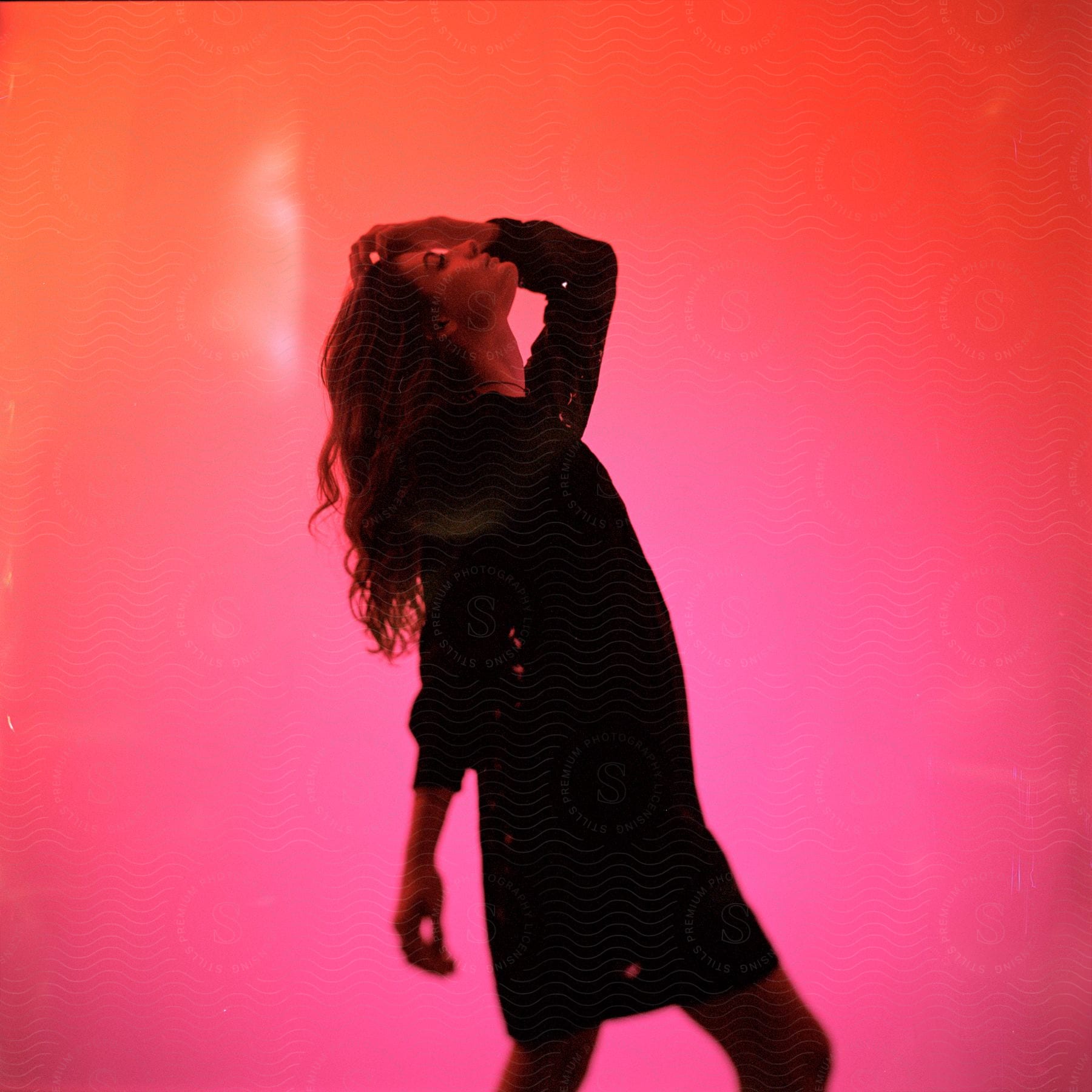 Young woman with one hand in her hair wearing a black dress against a pink background.