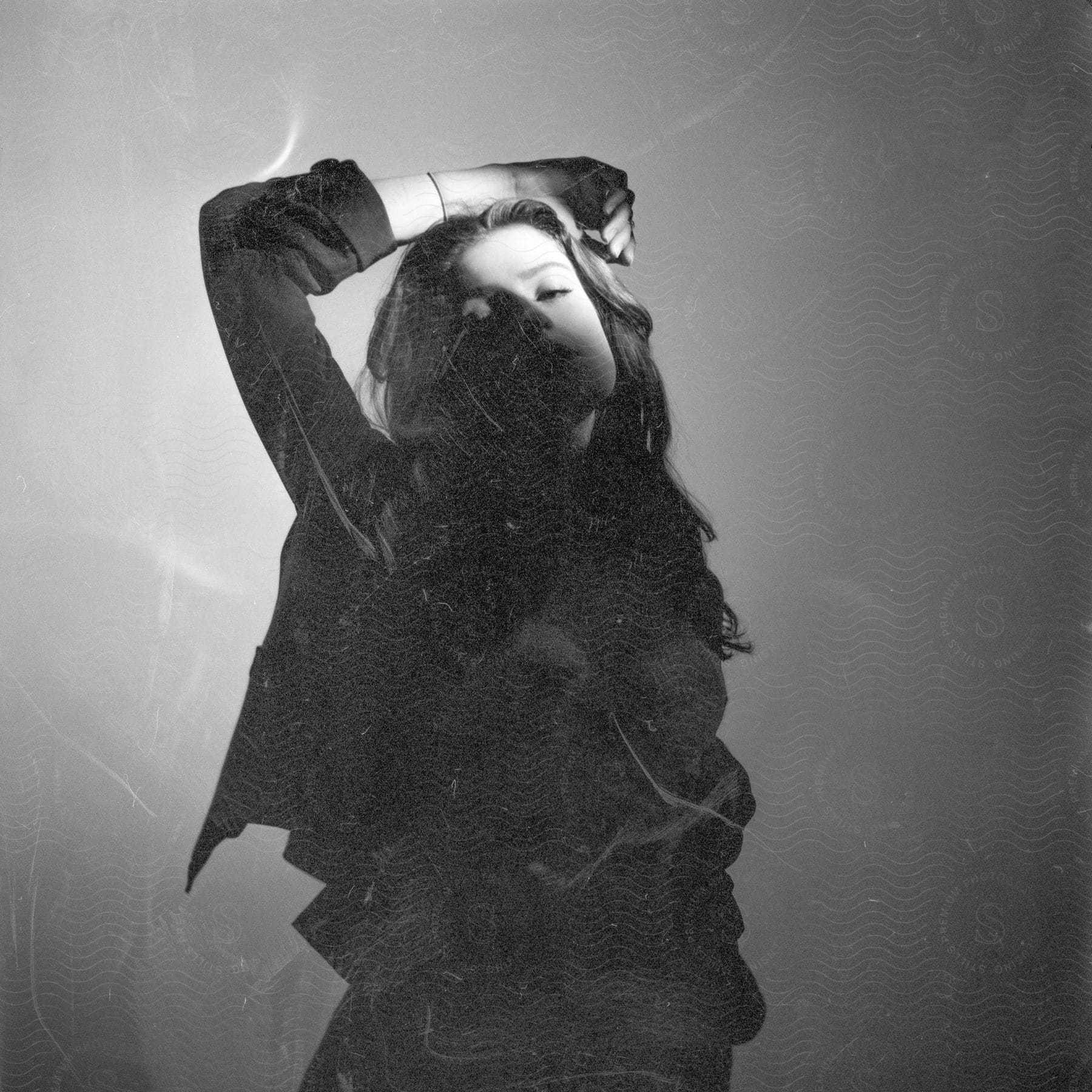 "A woman with wavy brown hair, wearing a black shirt, rests her arm on her head, half of her face bathed in soft light