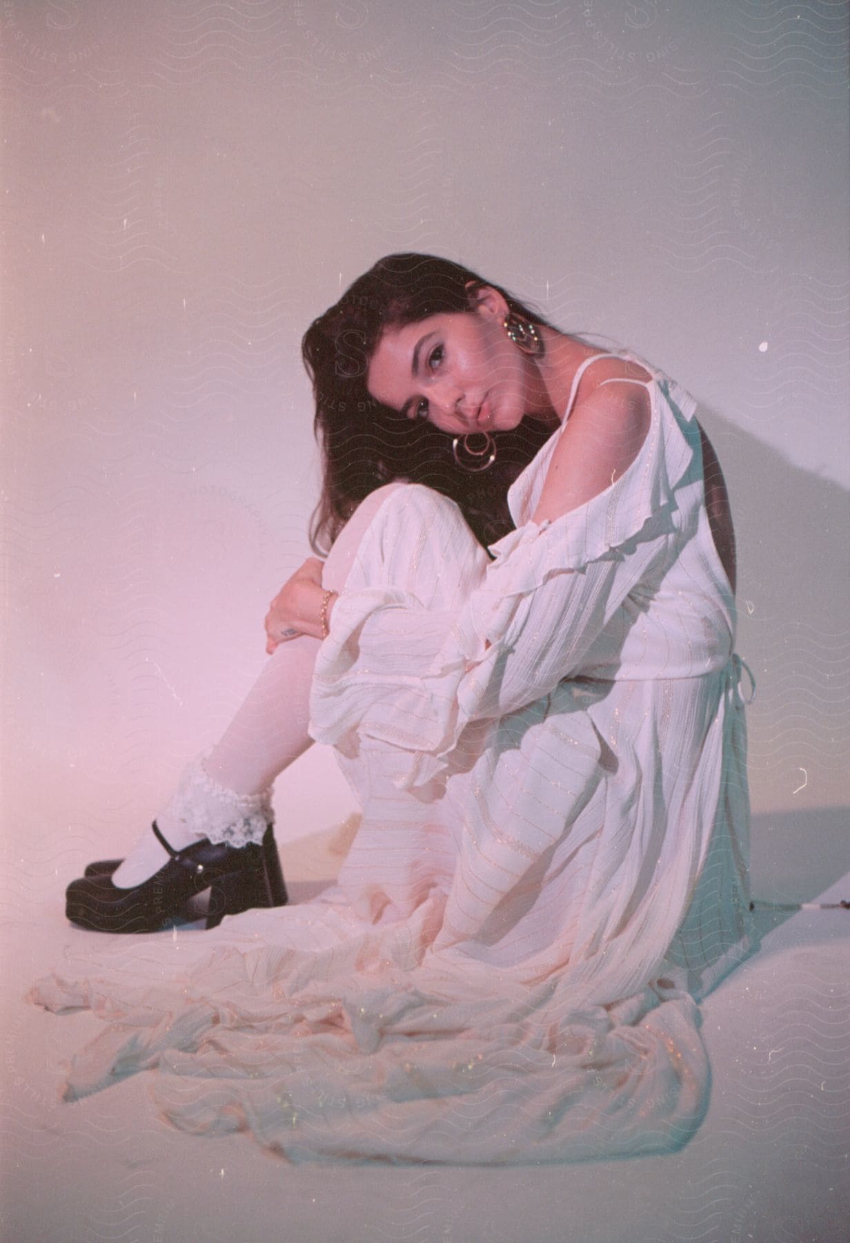 A young woman wearing a white dress and black heels sits on the floor with her head bent toward her knees.