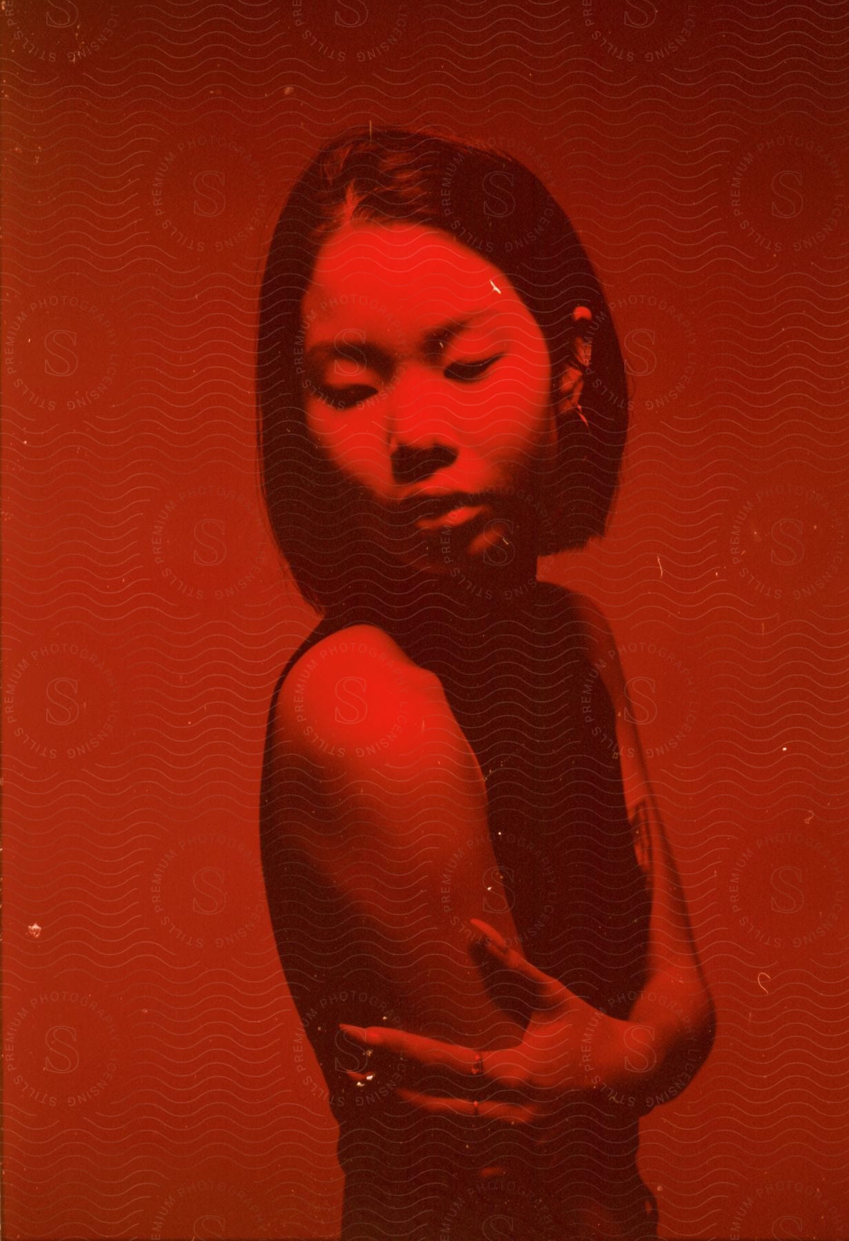 Woman with black top and straight hair stands arms crossed and looks over her shoulder in red lighting