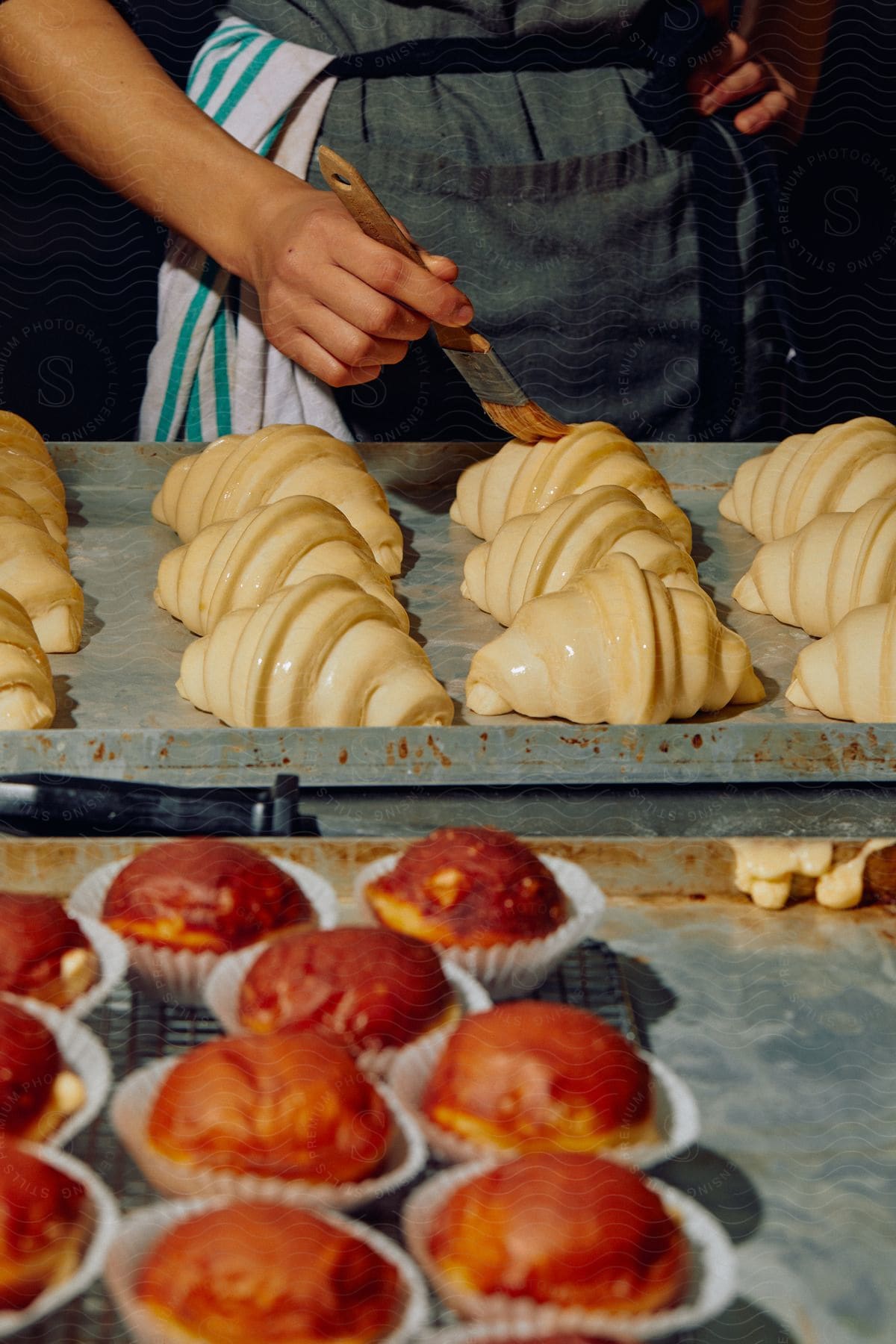 A baker in an apron brushes glaze on pastries while muffins cool in the foreground