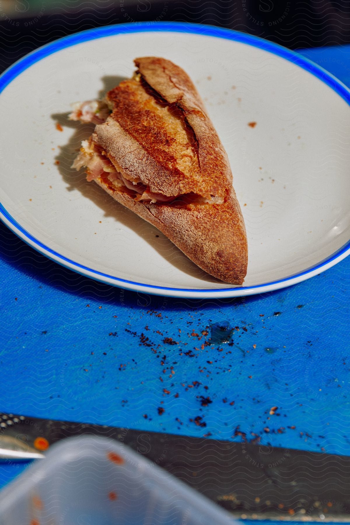 Half eaten sandwich on a plate with a knife container of food and crumbs on a table