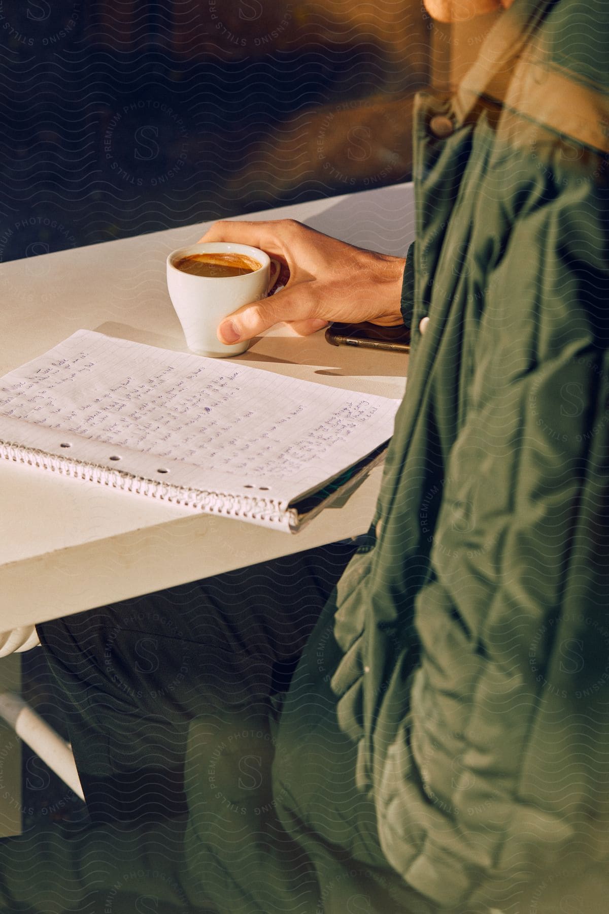 Stock photo of a man sitting at a table holding a cup of espresso while reading text from a notebook