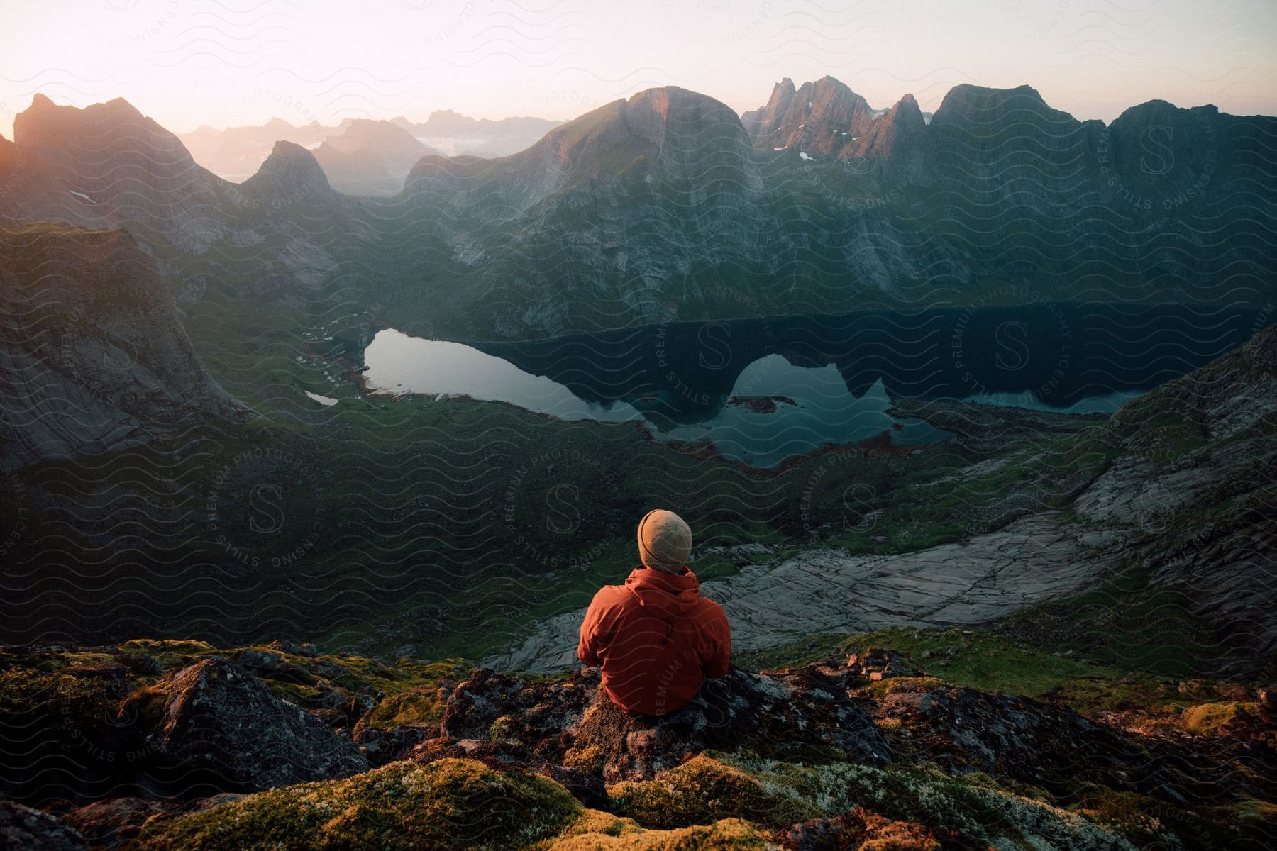 Stock photo of a person sits on a mountain top with other mountains and a lake before them