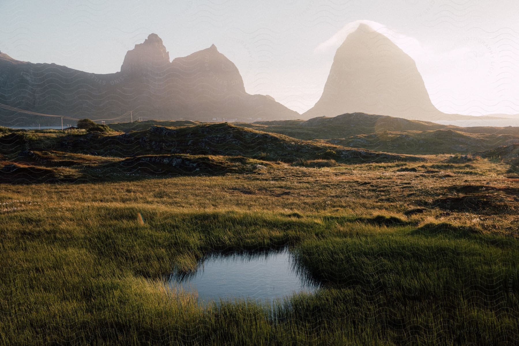 A small pond in front of tall mountain peaks with a road passing through on a hazy dawn