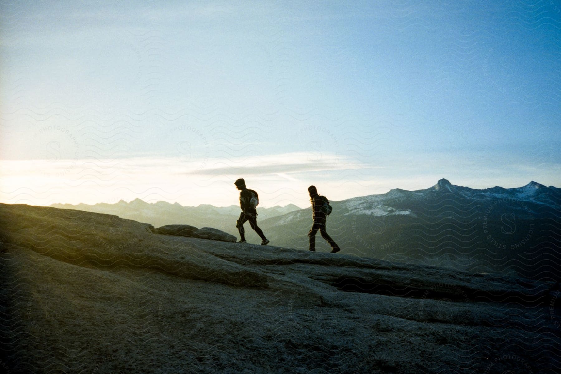 Two hikers walk on a snowcapped mountaintop at sunset with a snowcovered mountain range in the distance