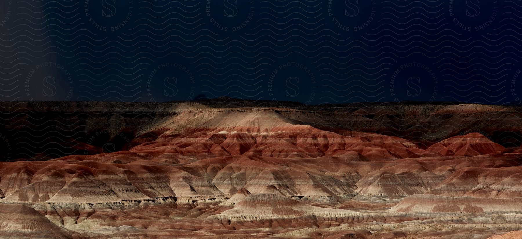 A bright light shines on painted desert hills under a dark night sky