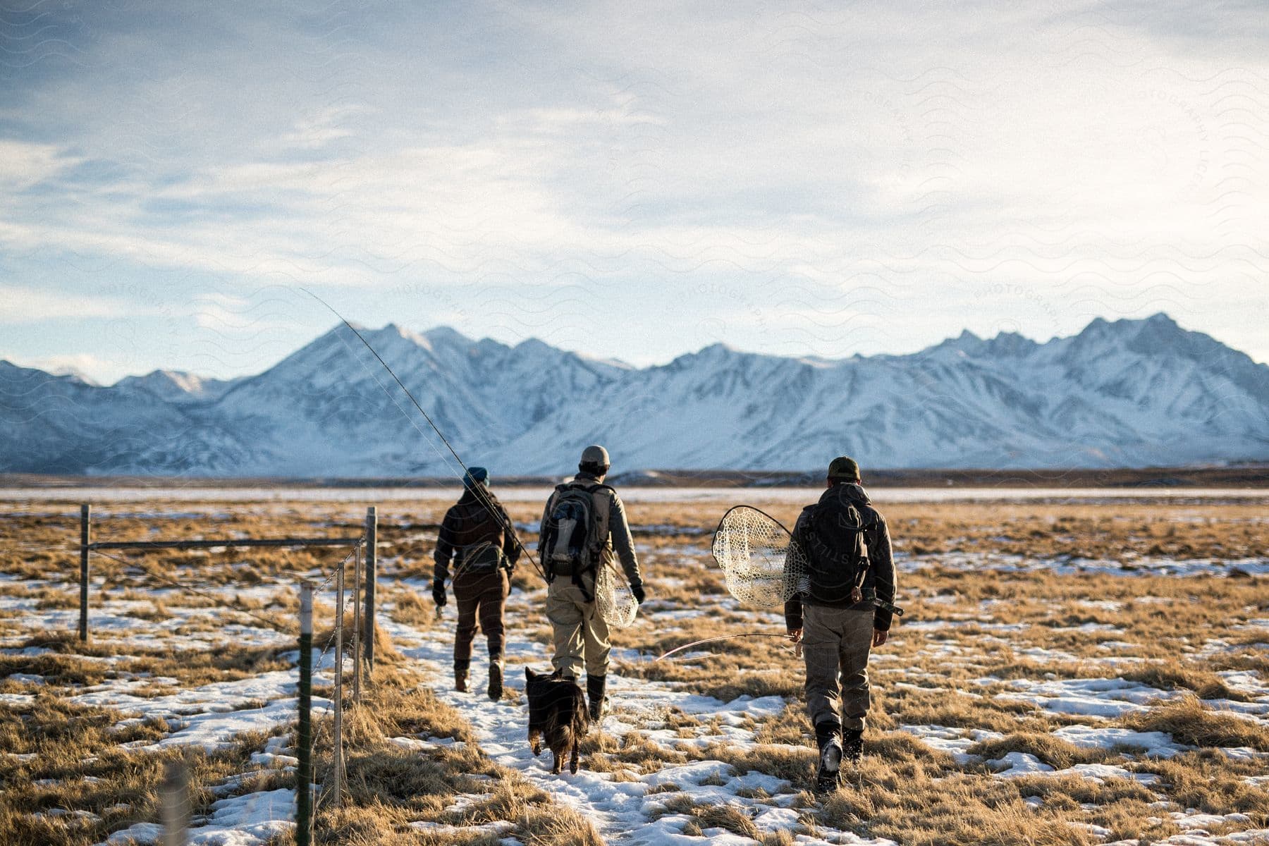 Three men with a dog walk across snow towards mountains on a cloudy day carrying fishing gear and backpacks