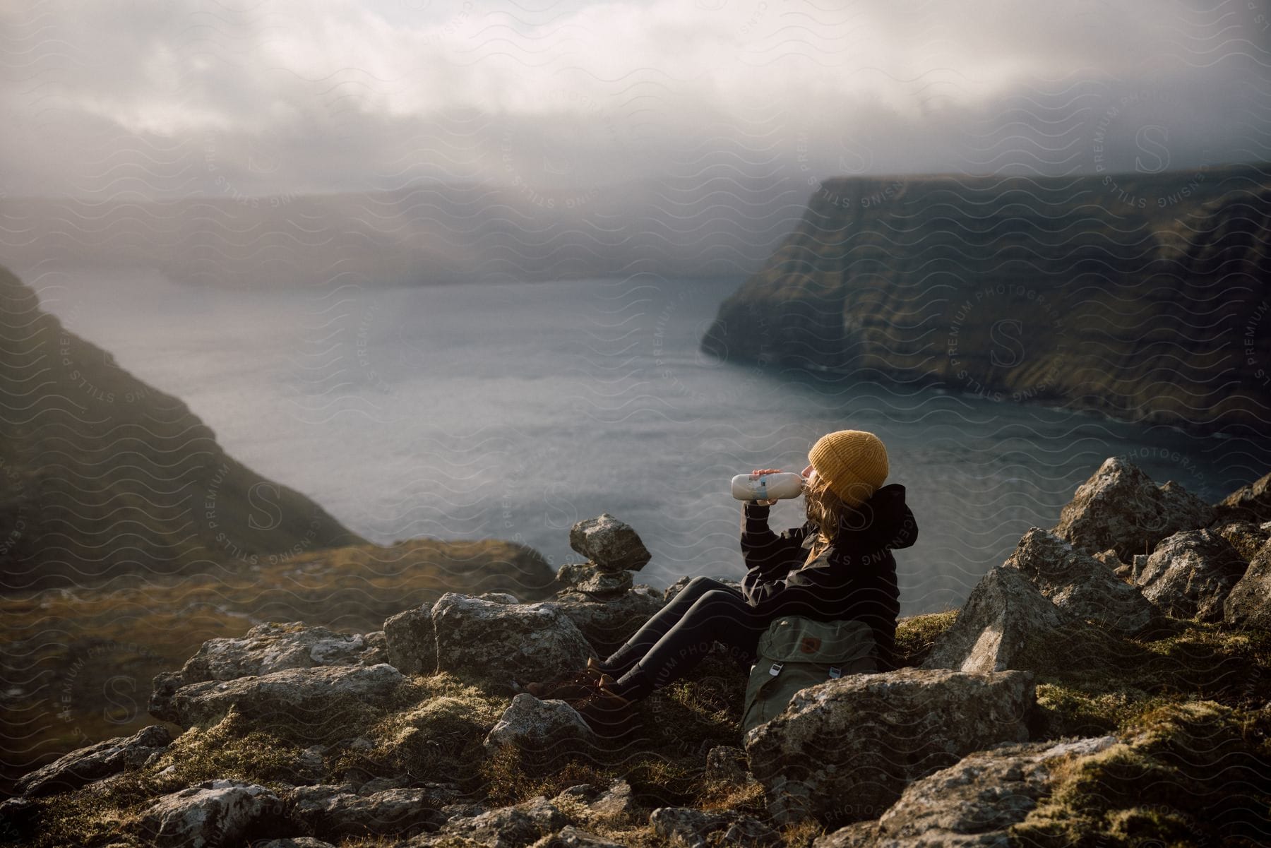 Woman sitting on rocks drinking from a bottle of water and enjoying the view of a lake surrounded by mountains.