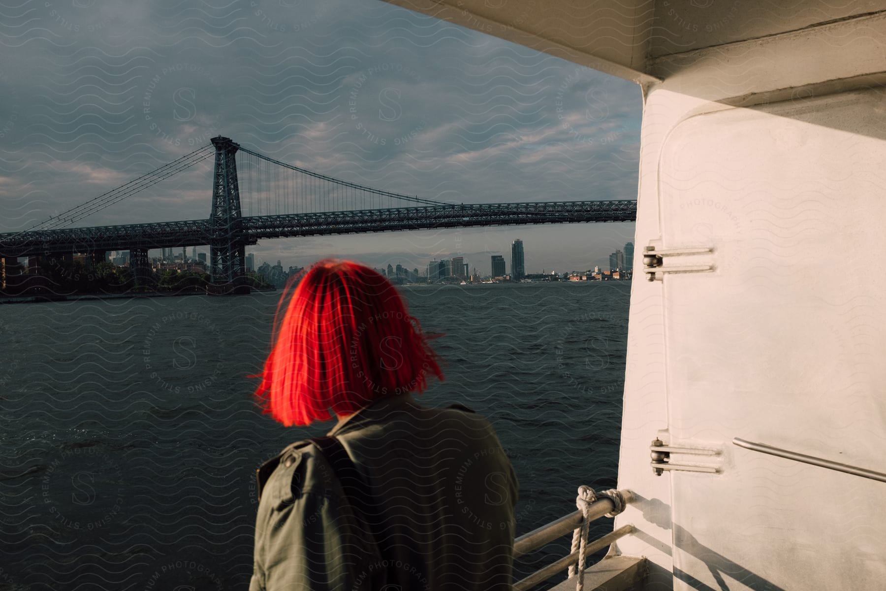 A woman in a green coat gazes at the water and a bridge from a boat on a cloudy day with a city in the distance