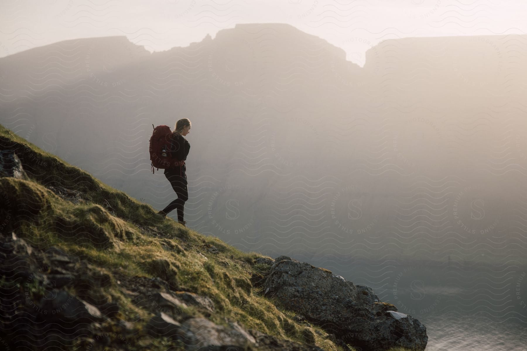 Hiker wearing a backpack walks down a mountain slope overlooking the water with mountains and fog in the distance