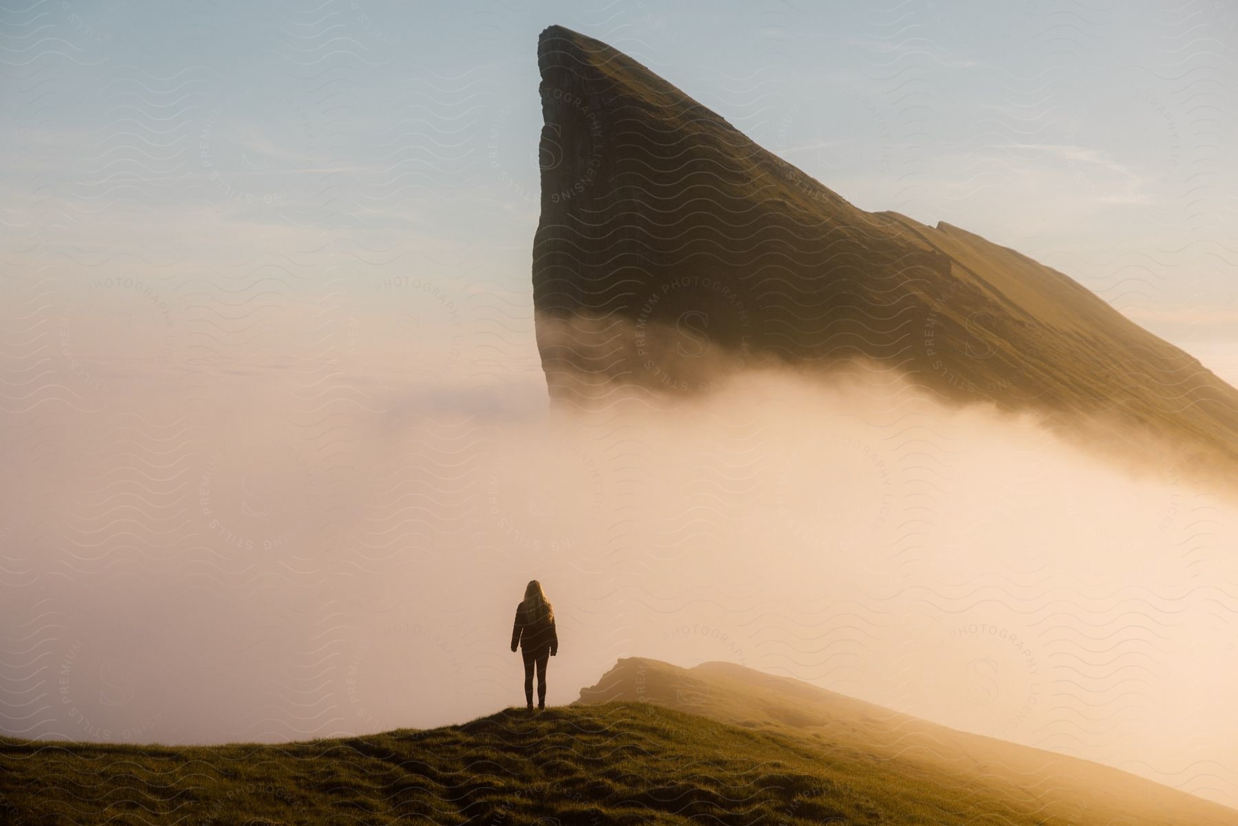 Woman standing on a hilltop with a rock formation protruding behind her in the outdoors at dusk or dawn with a mountain range and foggy landscape in the background