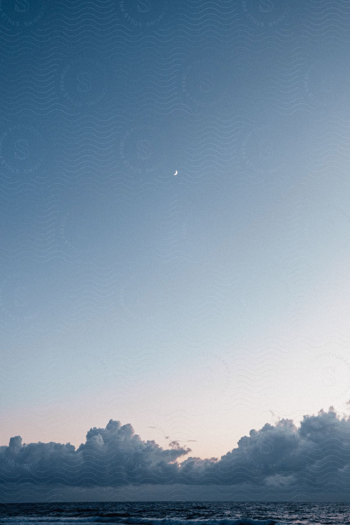 Clouds hang low over the water with a crescent moon in a blue sky