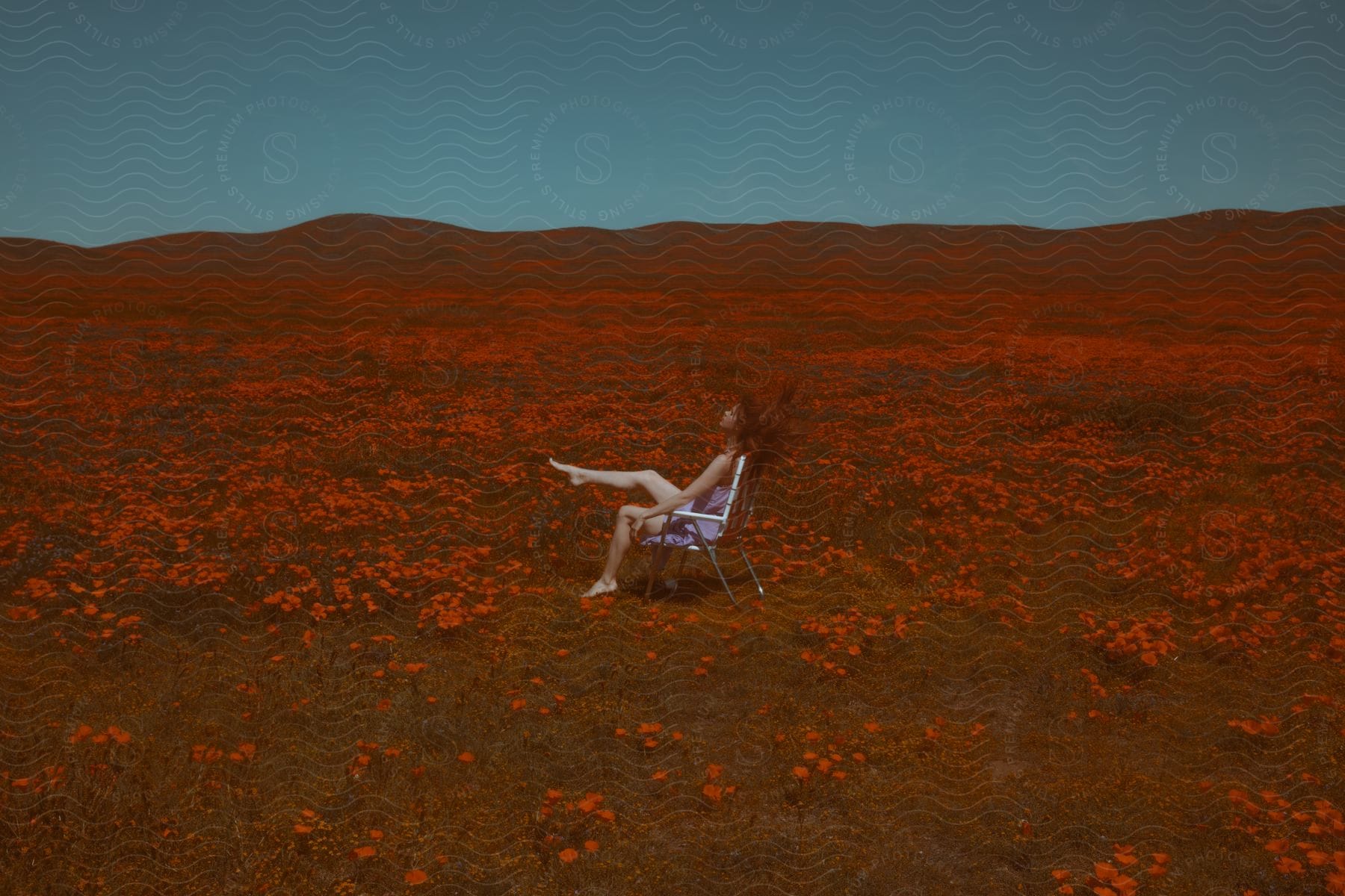 A young woman with red hair sits on a chair in a field of wildflowers kicking her leg up