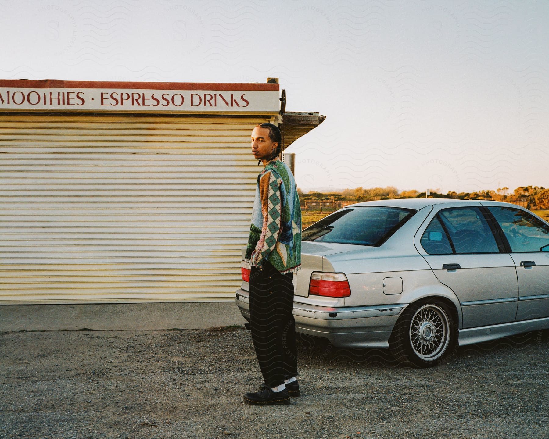 A man stands near his silver car wearing black pants and a green sweater