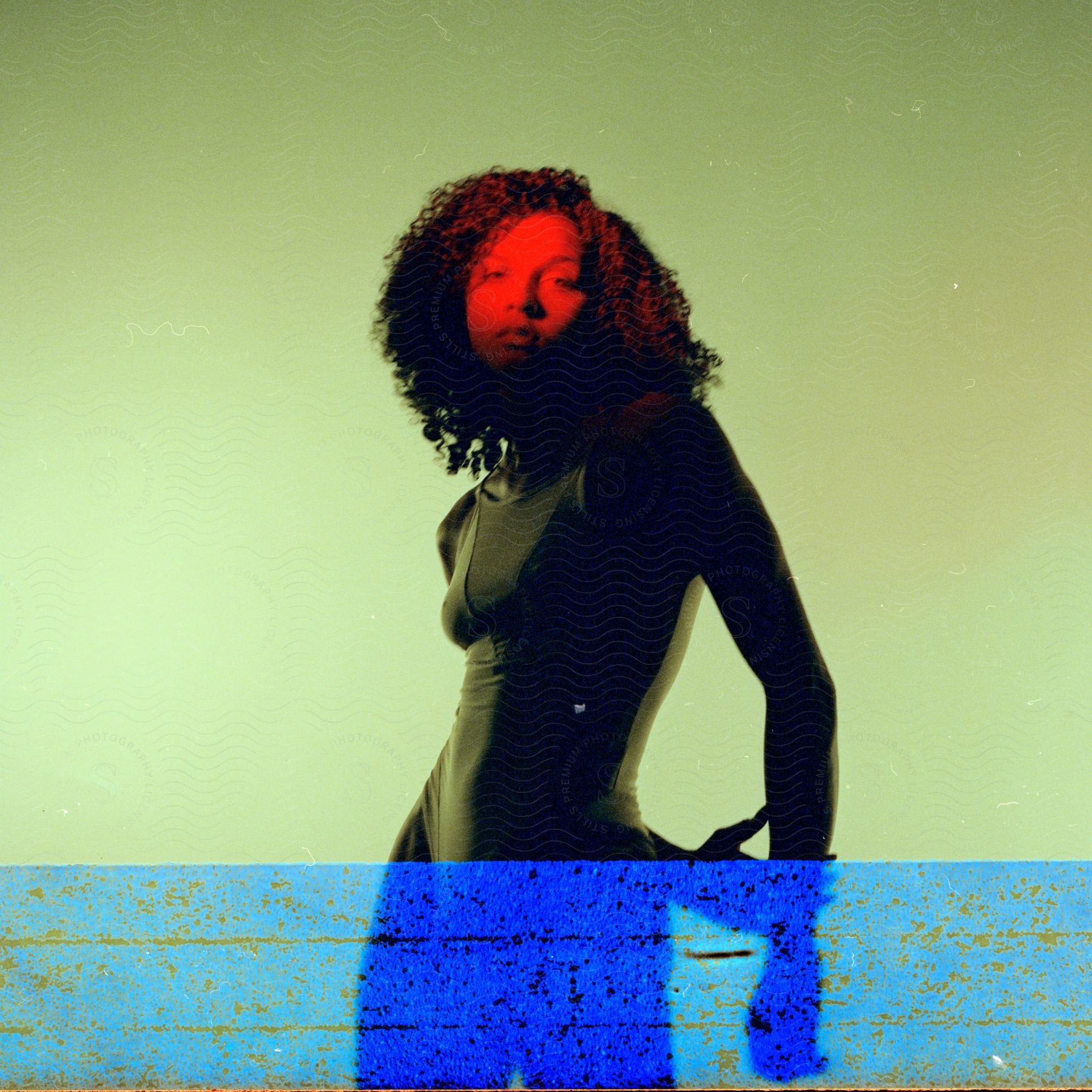 A woman with curly hair, clad in a yellow swimsuit, stands with her face bathed in red lighting