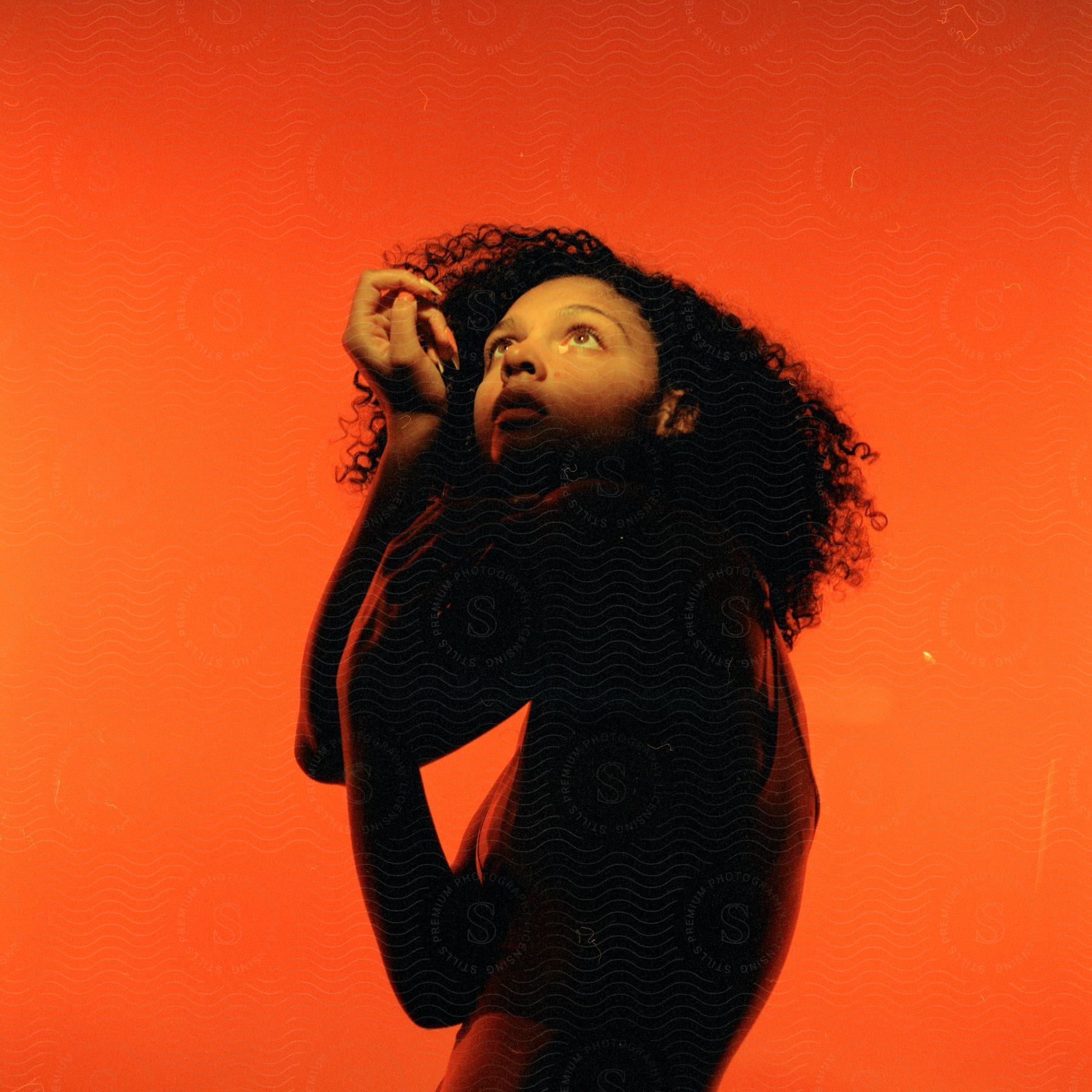 Woman with curly hair and a yellow swimsuit stands against an orange background, looking up