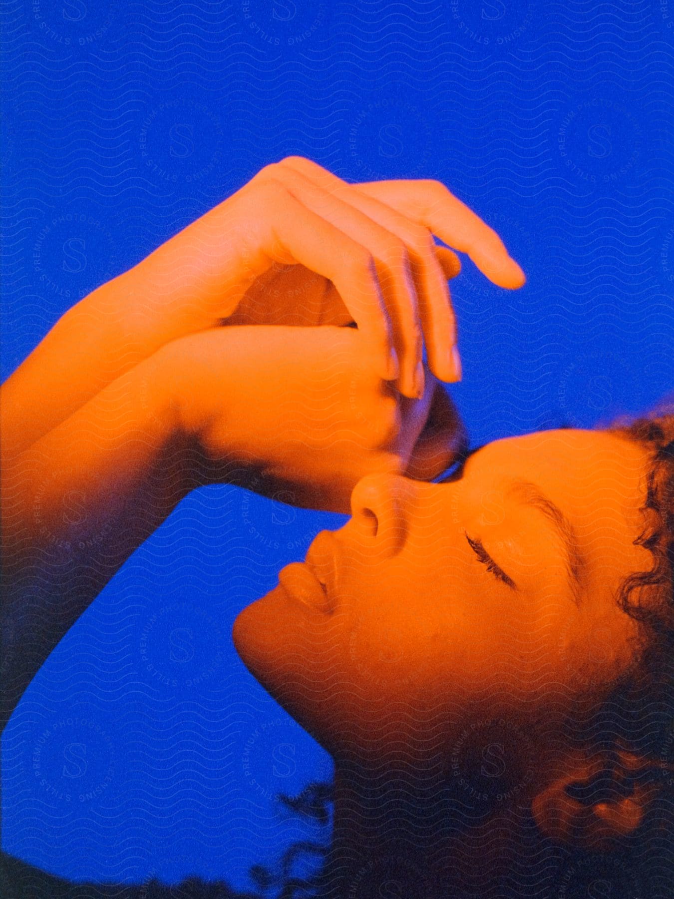 A female poses with her arms up above her face in orange lighting.