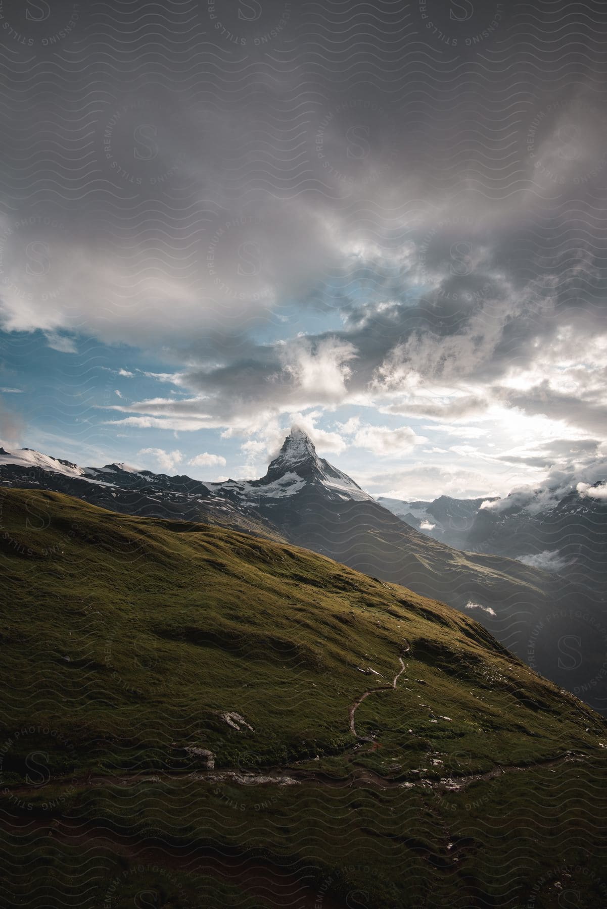 A grassy hill slope with a snowcapped mountain in the distance a path leading to the mountain under a cloudy sky and sun rays shining through the clouds on the right side