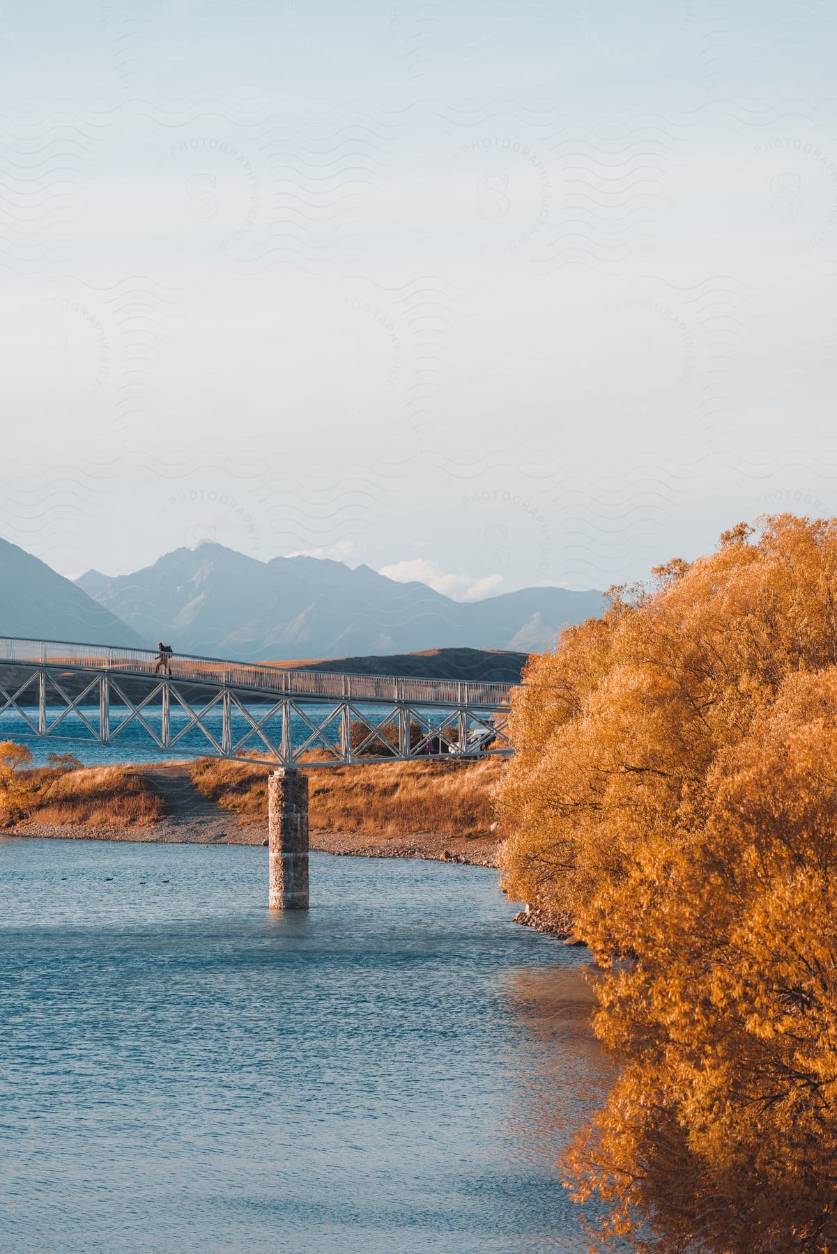 A white van parked by water a hiker crossing a bridge over a lake golden brown leaves on water bank trees and a cloudy day time sky in new zealand