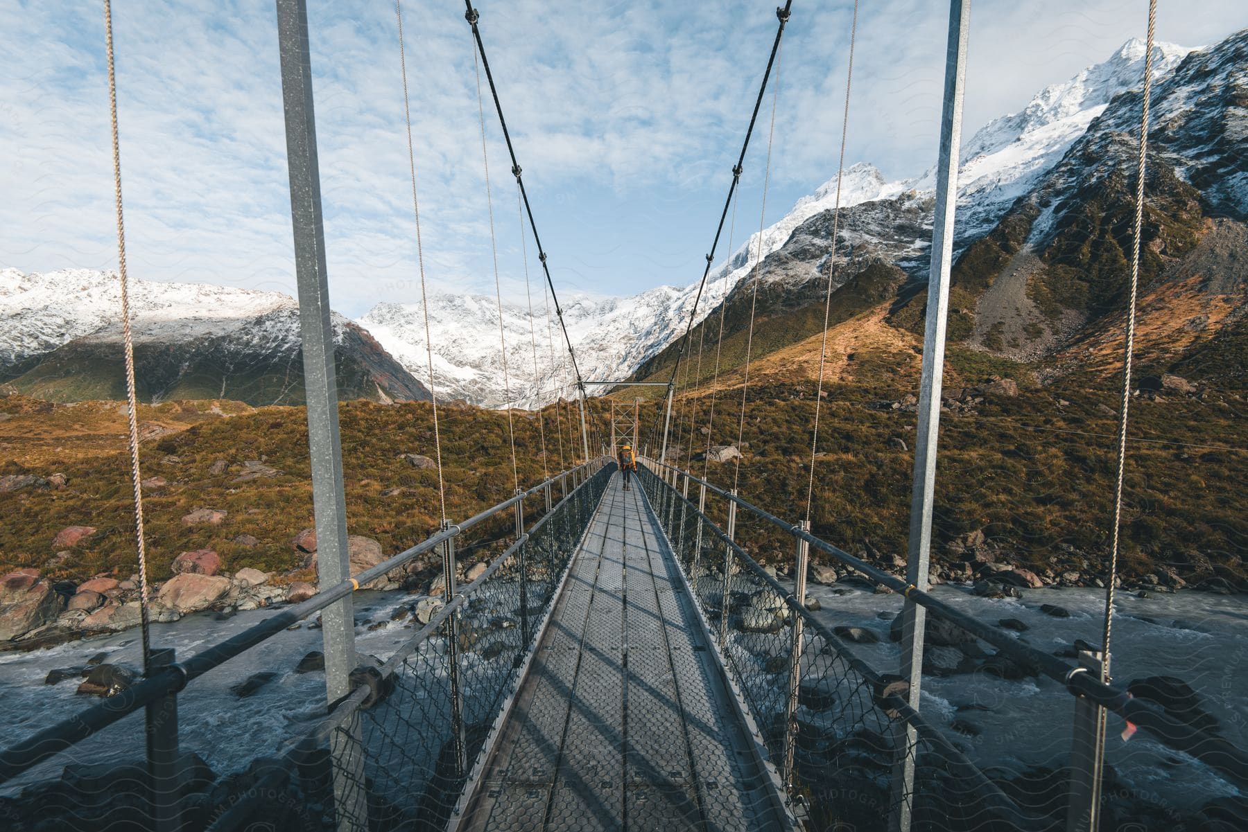 A hiker walks across a suspension bridge over a rocky river with a snowcapped mountain in the background and green hills on the other side