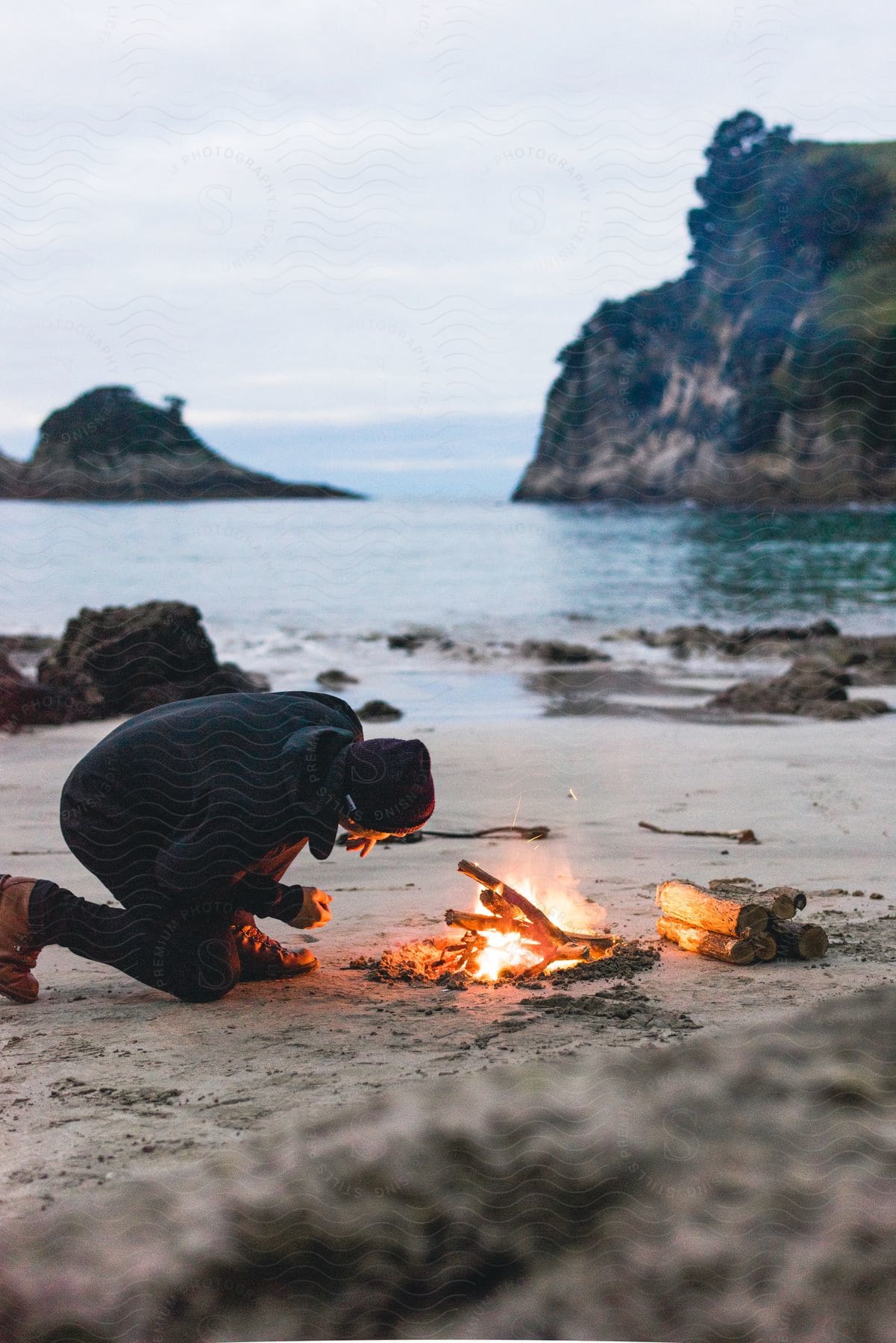 A man setting up a beach bonfire at sunset with mountains in the background