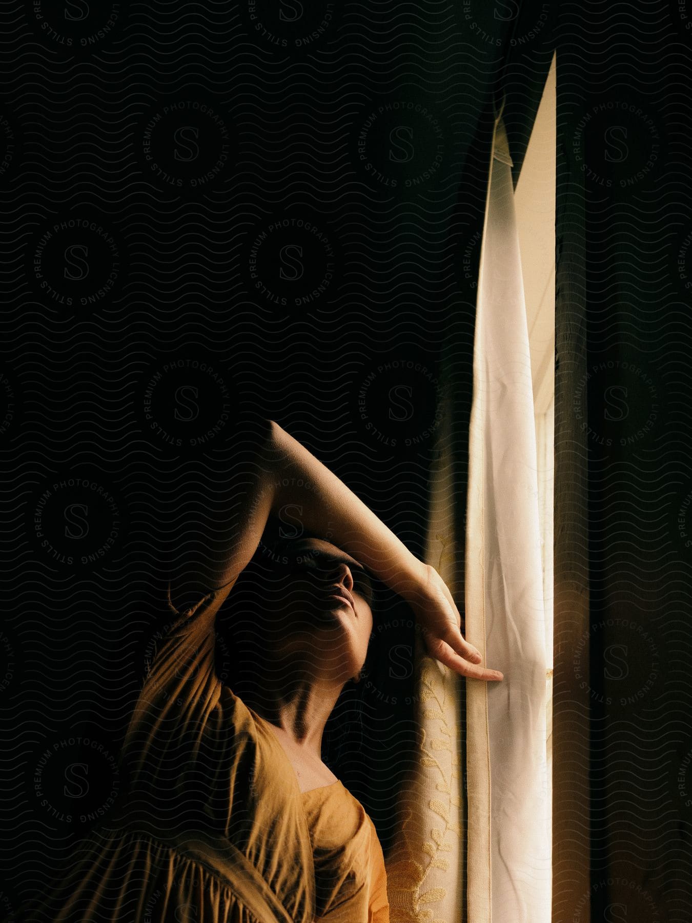 A darkhaired woman with her arm over her face stands next to a window