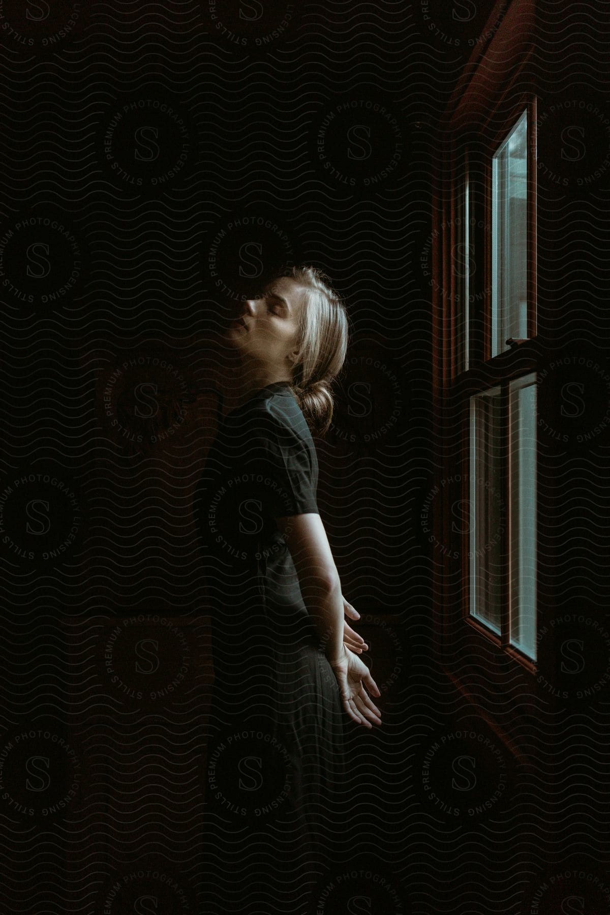 Blond woman in brown dress stands with head tilted up eyes closed hands behind her back in front of window with sun shining through cloudy sky