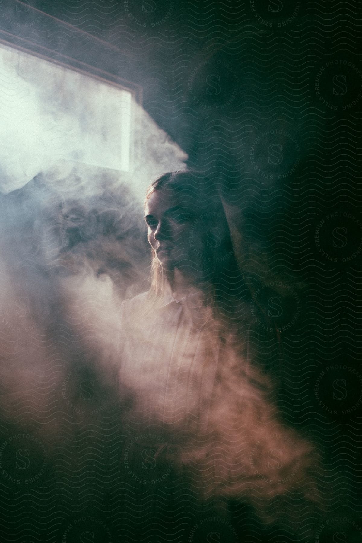 A woman standing next to a window in a smoky room with natural light coming in