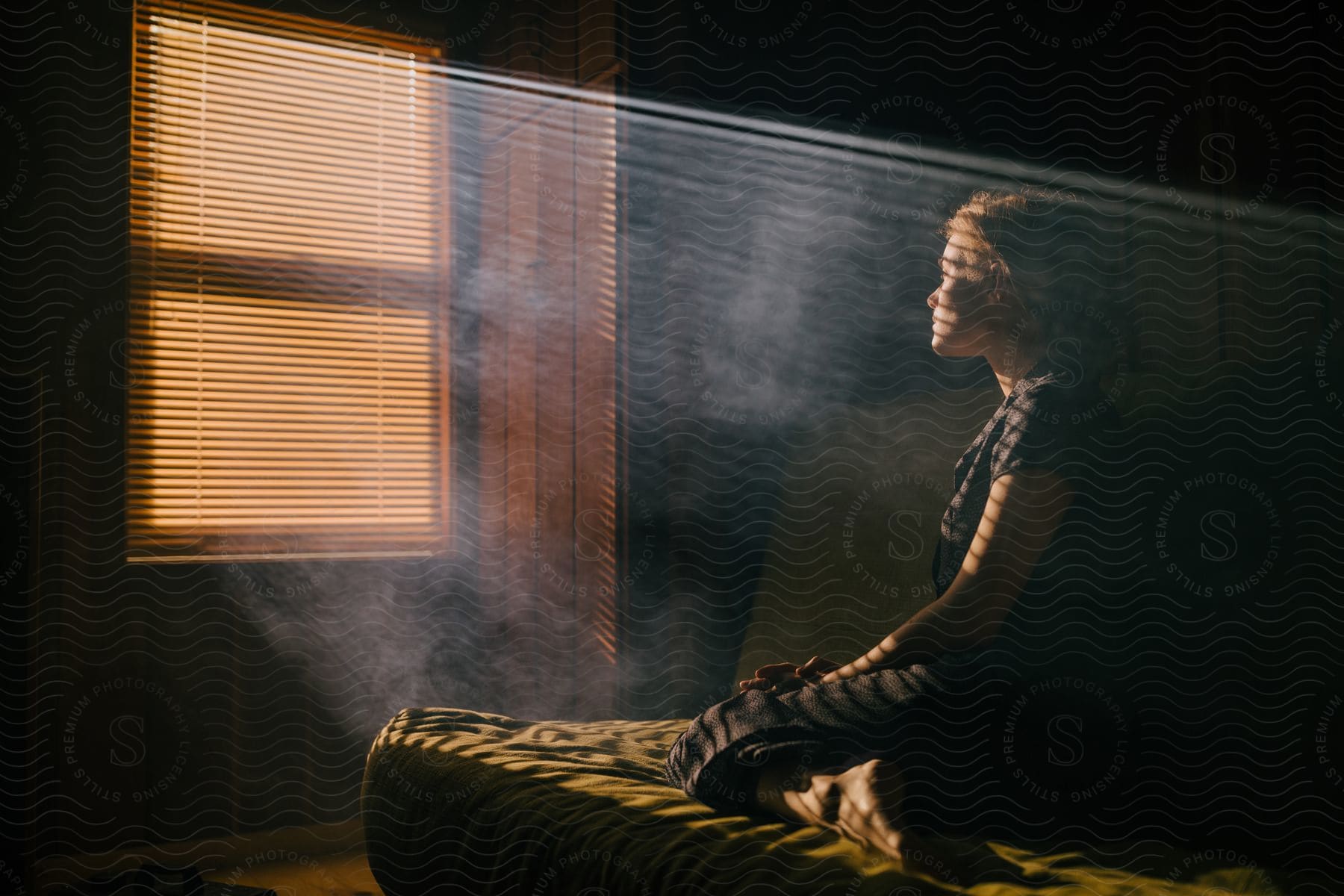 A woman sitting on a bed facing a window with shaped light covering her