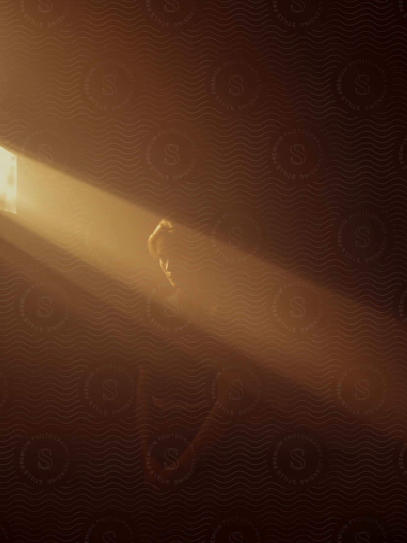 Blonde Woman Stands In Dark Room While Sun Streaming Through Narrow Window