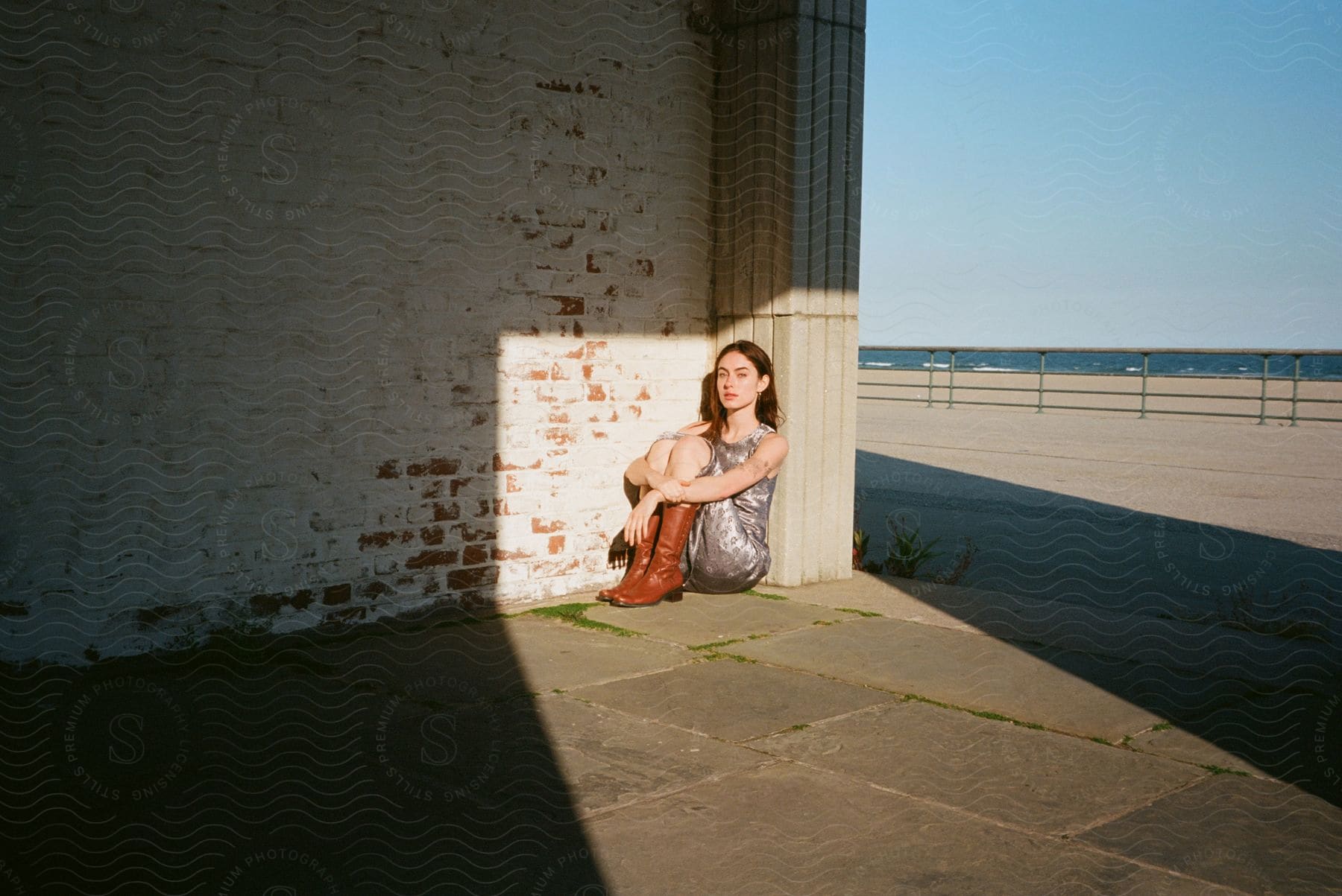 Woman sitting with knees to chest and arms wrapped around them leaning against stone pillar next to brick wall near the ocean