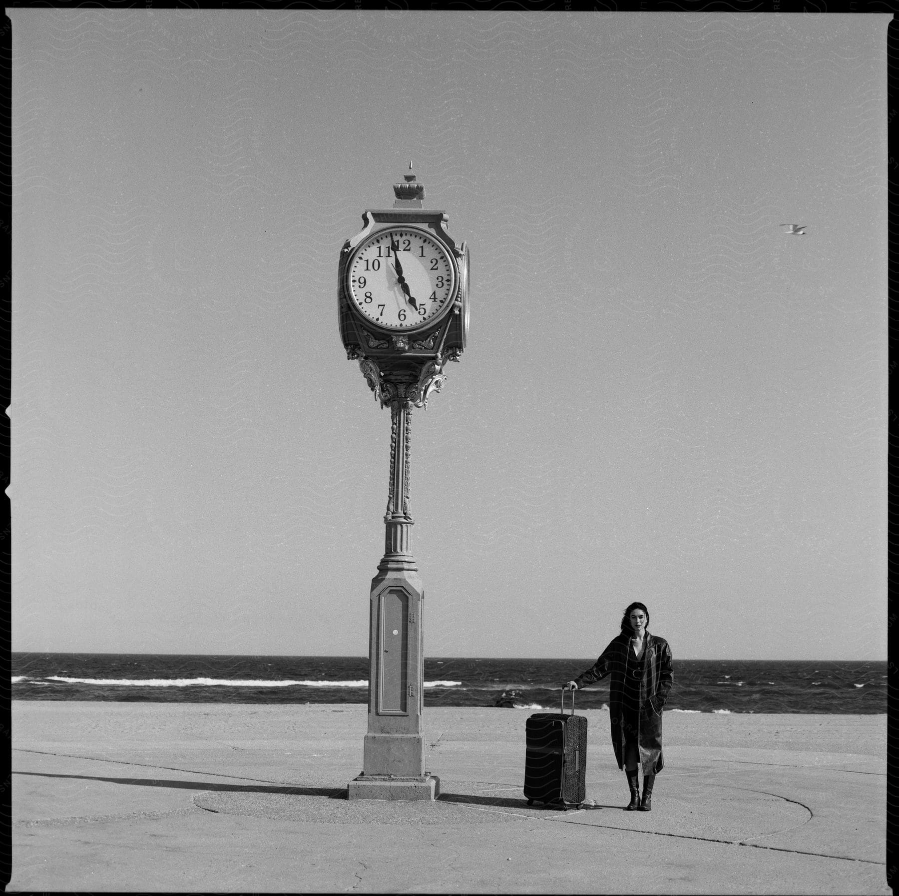 Stock photo of a woman in a trench coat with a suitcase near a clock on the beach
