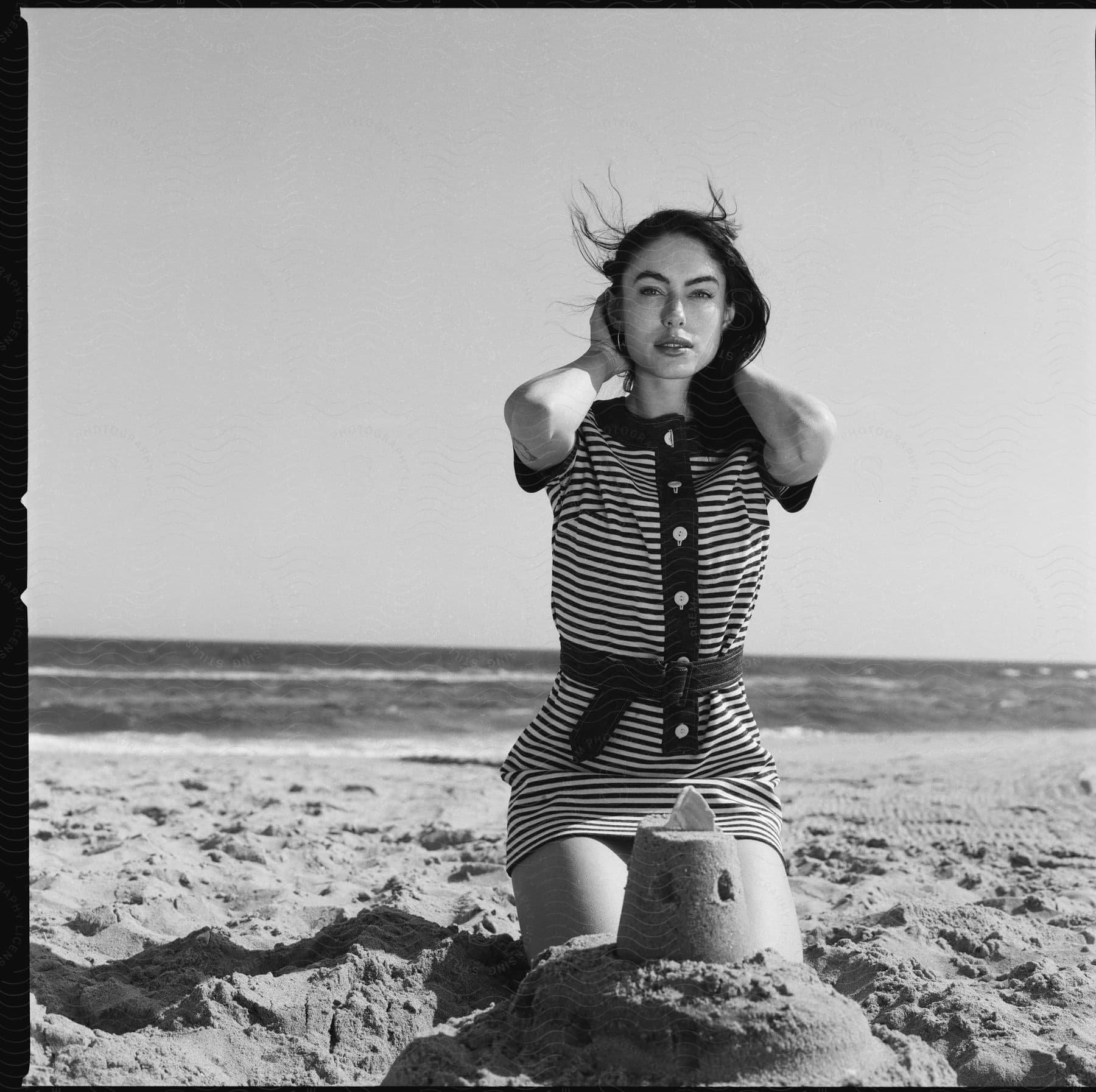 A white woman gracefully poses kneeling before a sandcastle at the beach exuding serenity and relaxation